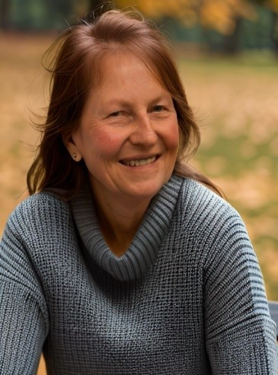 Eine lachende Frau mit braunen Haaren, trägt einen grauen Pullover, im Hintergrund sind Herbstblätter und Bäume.