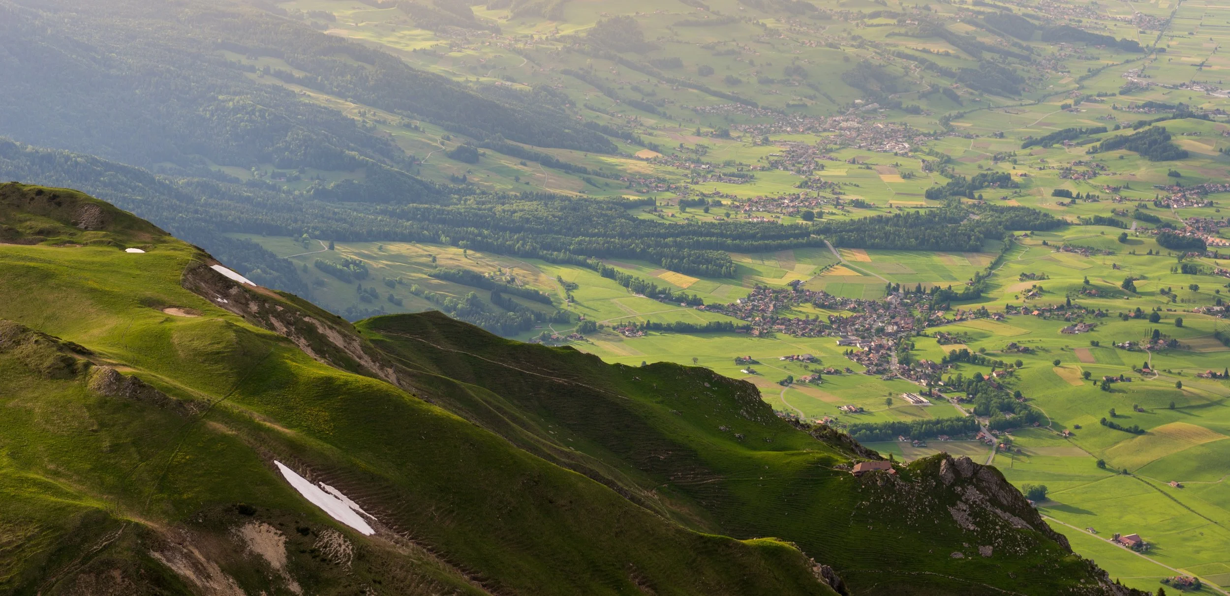 Blick von einem bergigen Gebiet auf eine grüne tiefliegende Landschaft mit Wiesen, Wäldern und kleinen Dörfern.