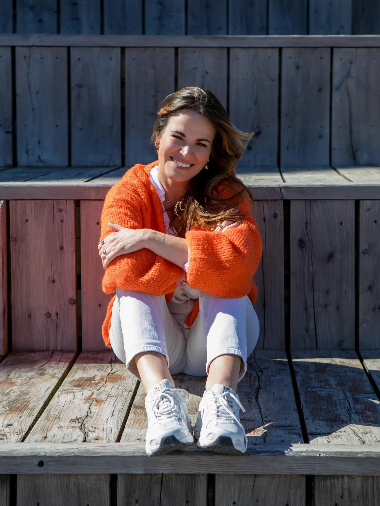 Hayley Buchan Teen Resilience Coach with brown hair, wearing an orange sweater and white pants, sitting on wooden steps at Stage@TheDock in Hull, smiling with her arms crossed, in front of a wooden fence outdoors.