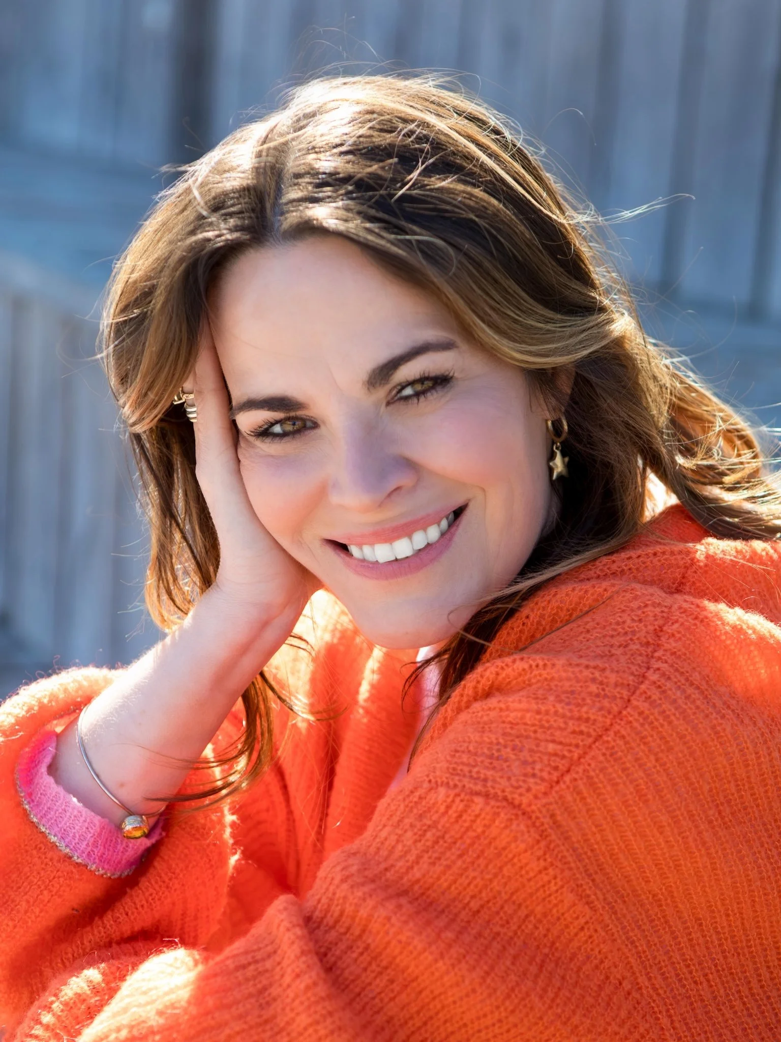 Close-up of Hayley Buchan Teen Resilience Coach with brown hair, smiling and touching her face, wearing a bright orange cardigan and jewellery.