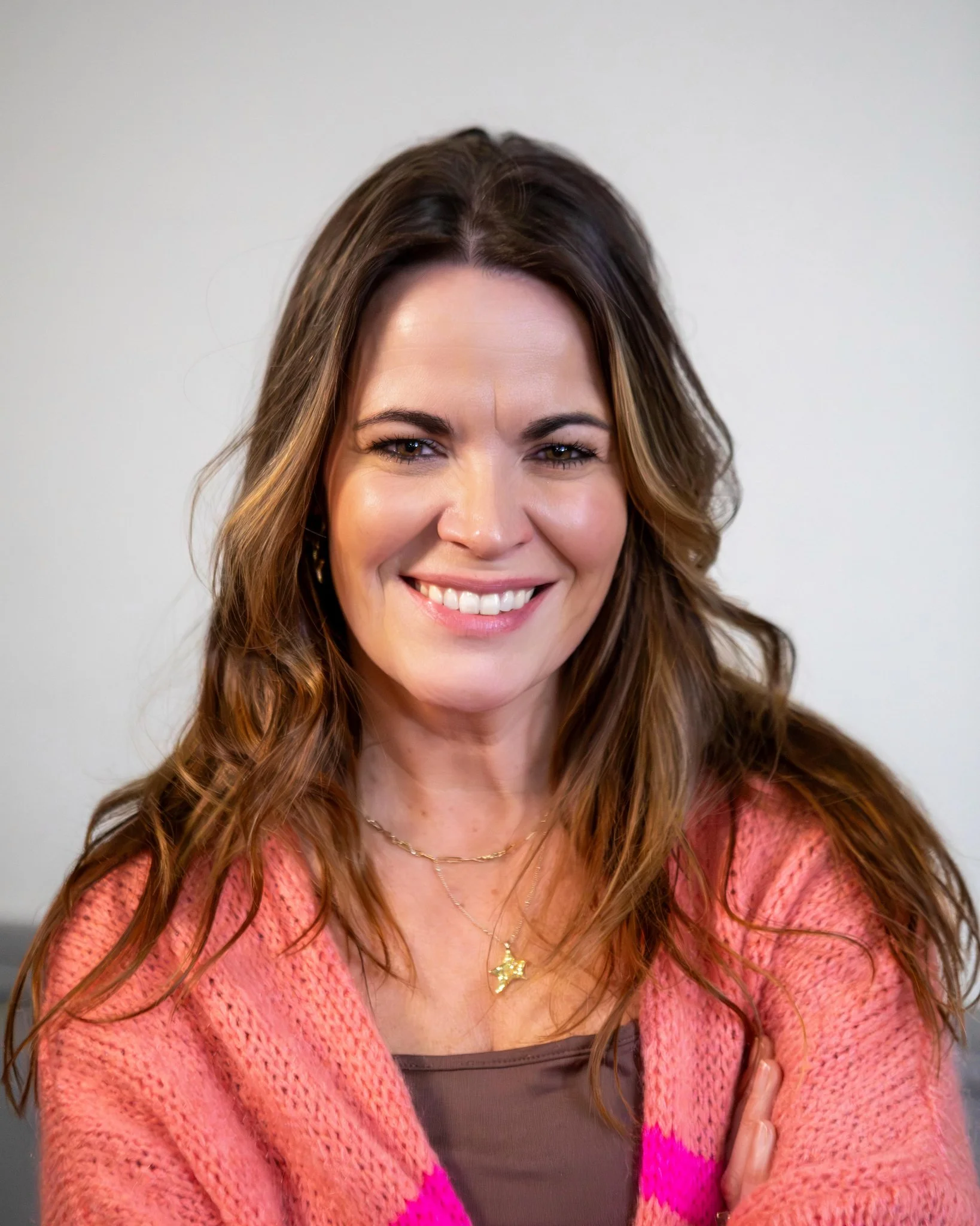 Close-up of Teen Resilience Coach Hayley Buchan, smiling with wavy brown hair, wearing a pink knitted cardigan, gold star necklace, and a dark top underneath, against a plain white background.