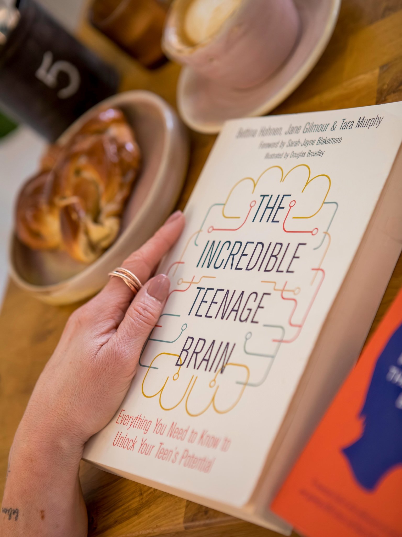 Hayley Buchan Teen Resilience Coach holding a book titled "The Incredible Teenager Brain" with a hand showing a gold ring. The background shows a plate of pretzels, a cup, and a box of popcorn, all on a wooden table.