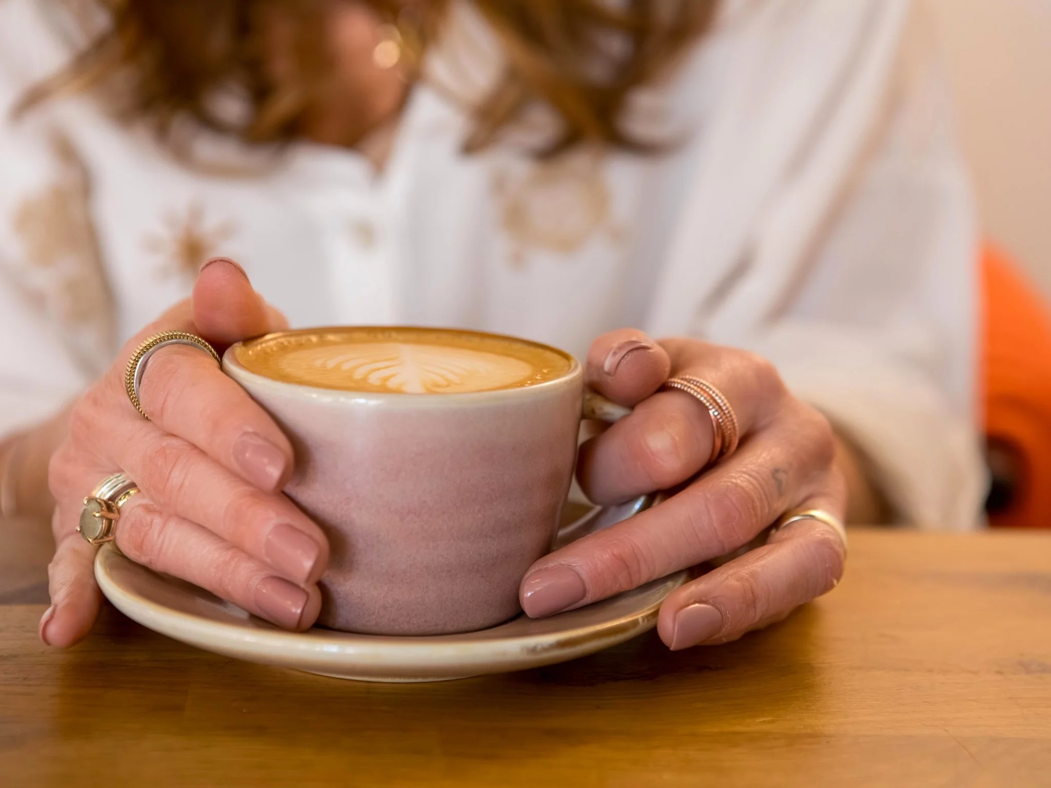 Hayley Buchan Teen Resilience Coach holding a cappuccino with latte art in a ceramic cup and saucer, wearing rings and a white shirt, sitting at a wooden table at Flour & Feast Bakery, Hull, Humber Street, Fruit Market.