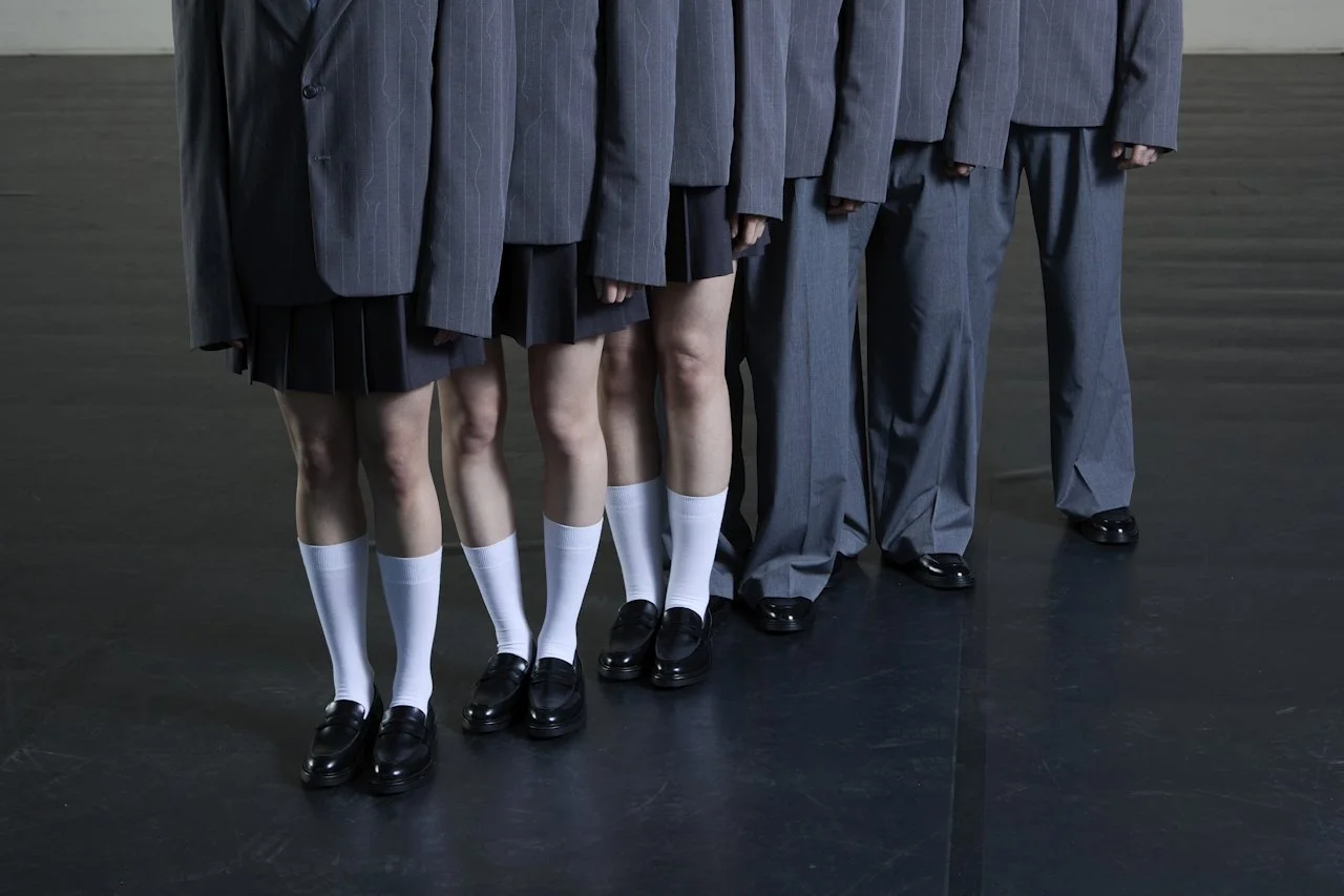 A line of five students dressed in school uniforms, with three girls wearing skirts, white socks, and loafers, and two boys wearing gray suits, all standing on a dark floor.