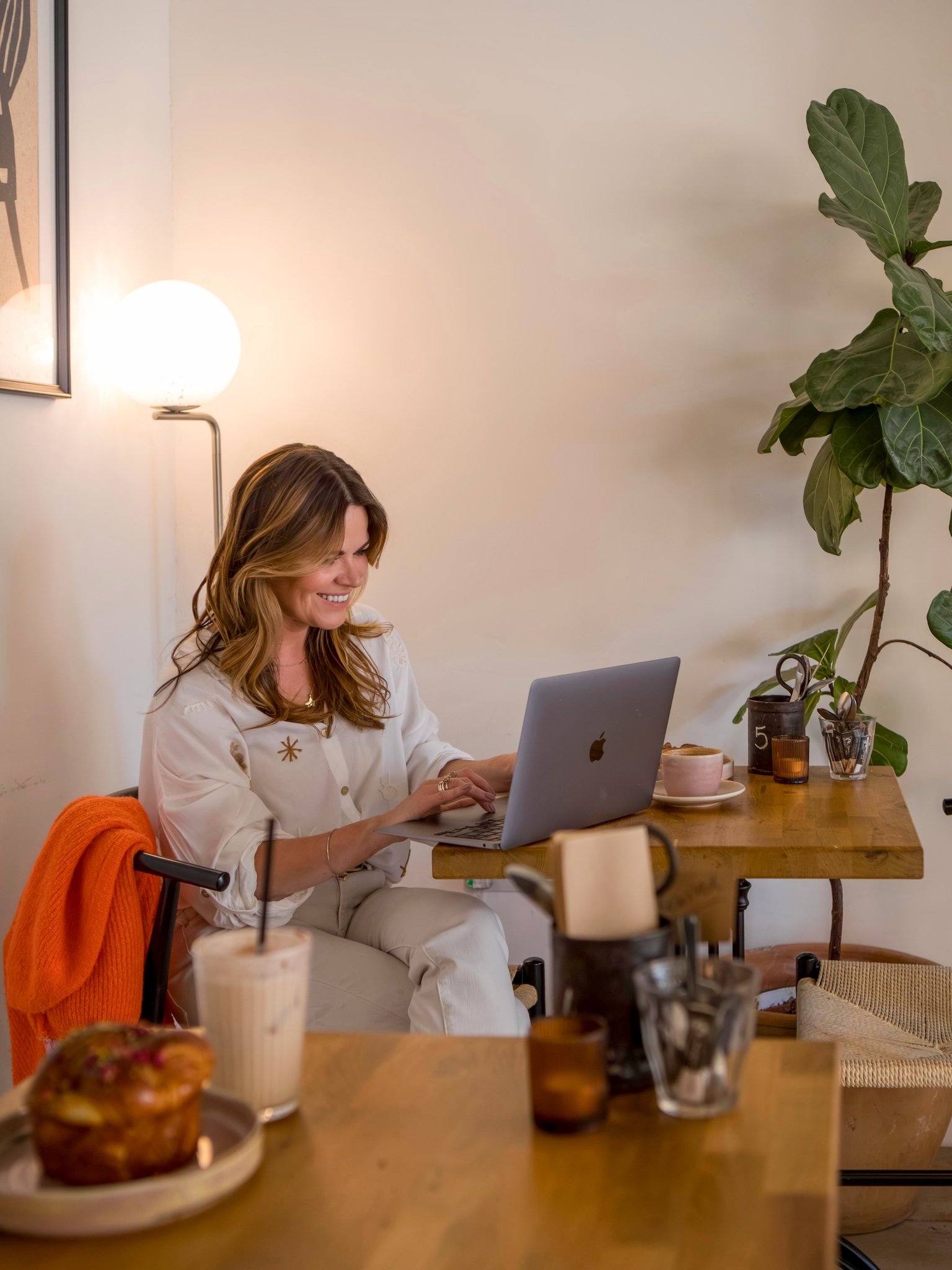 Hayley Buchan Teen Resilience Coach, with long hair sitting at a wooden table working on a laptop in a cozy, modern room with a large green plant, a lamp, and various drinks and food on the table at Flour & Feast Bakery on Humber Street in Hull.