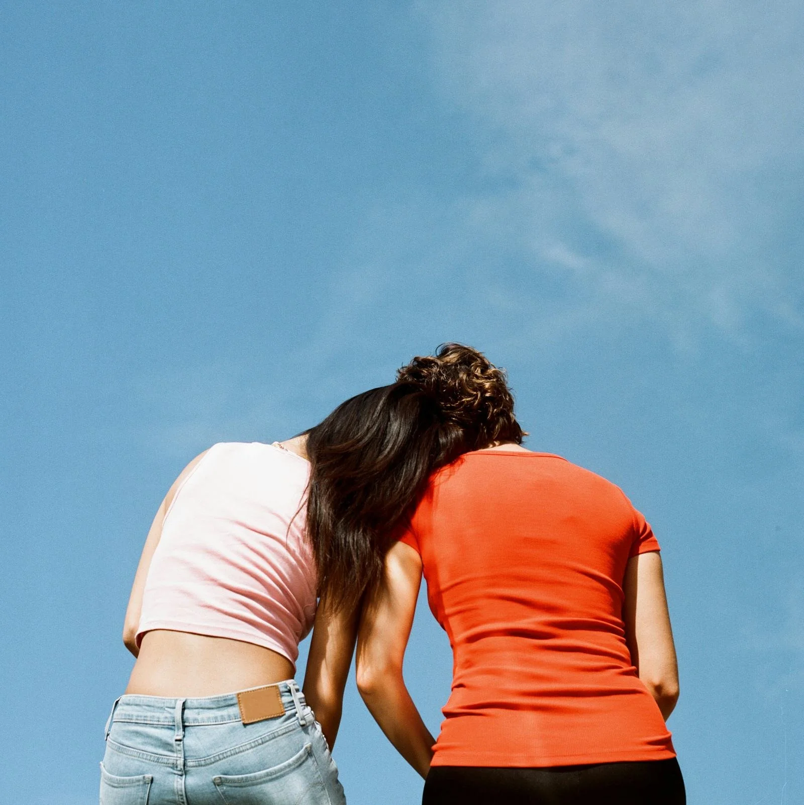 Two women with curly hair are hugging each other outdoors against a clear blue sky. One woman is wearing a pink top and light blue jeans, and the other is wearing a red top.