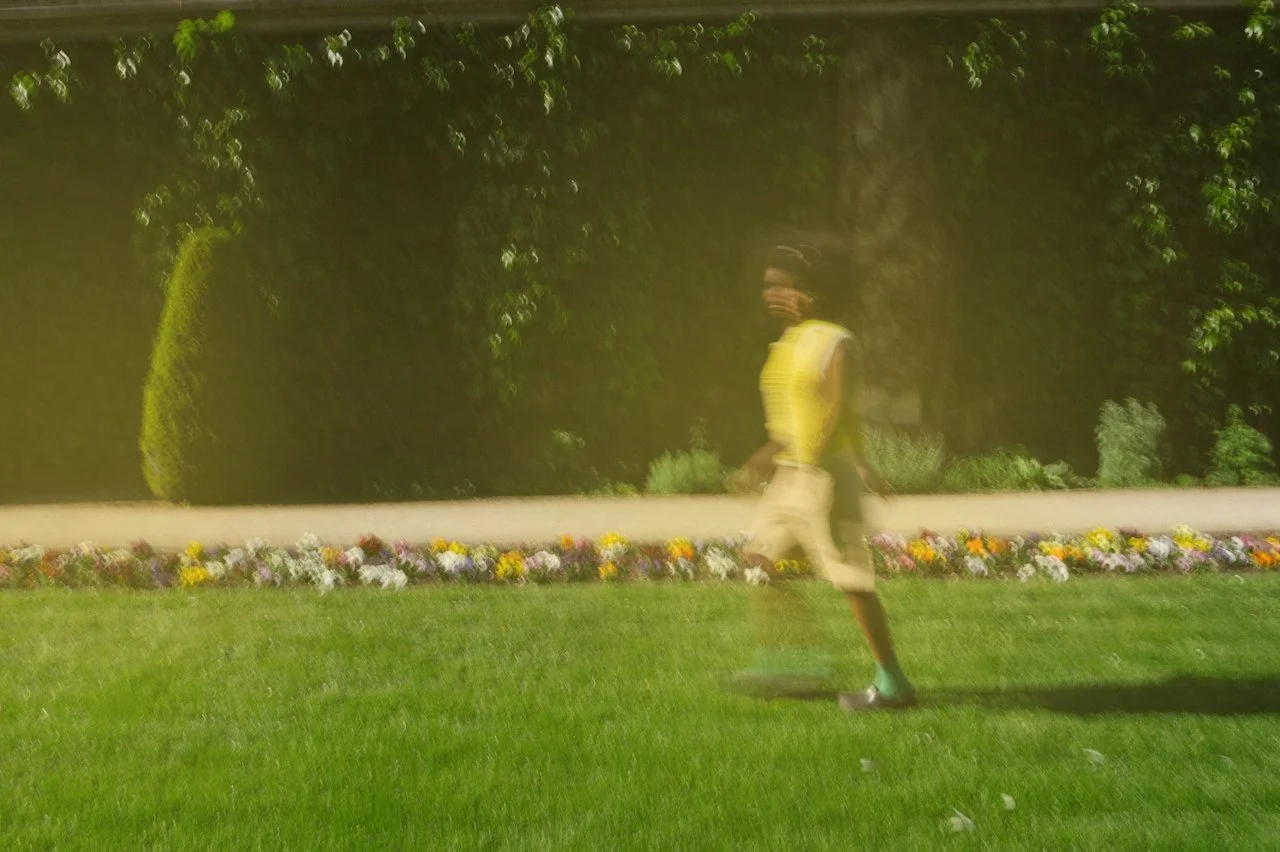 A person is walking outdoors on a grassy area near a flower bed, wearing a yellow top and light-colored shorts, with trees in the background.