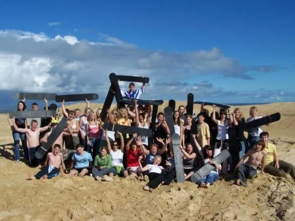 Archbishop Sentamu Academy teachers and students posing with large black foam sticks on sandy Australian beach under a blue sky, on a school trip organised by Teen Resilience Coach, Hayley Buchan.