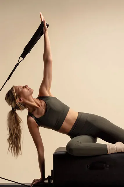 A woman performing a stretch or exercise on a padded bench, reaching upward with one arm while supporting herself with the other arm on the bench.