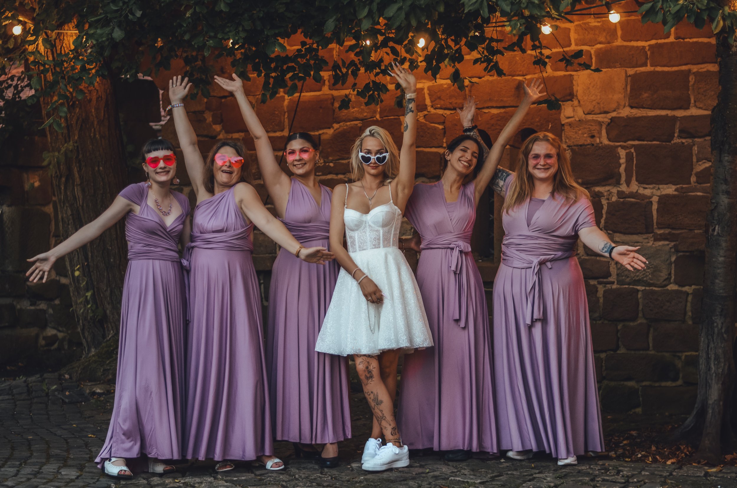 Sechs Brautjungfern / Trauzeuginnen in lilafarbenen Kleidern und die Braut in einem weißen Kleid posieren glücklich beim Feiern im Freien bei Nacht, umgeben von Bäumen und Steinmauer.