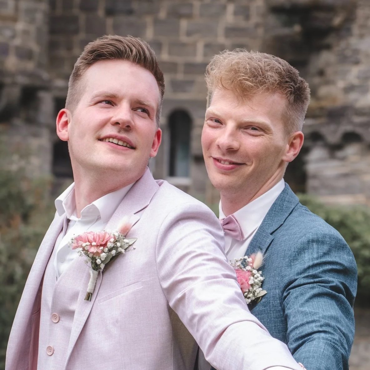 Zwei Männer in Anzügen, die sich zur Hochzeit schmücken, mit Blumen auf der Brust, vor einer alten Steinmauer.