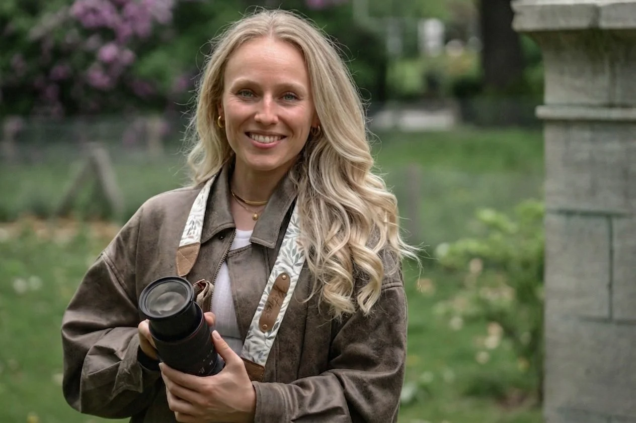 Eine lachende Frau mit langen blond lockigen Haaren, die eine Kamera hält, steht im Grünen, im Hintergrund blühende Bäume.