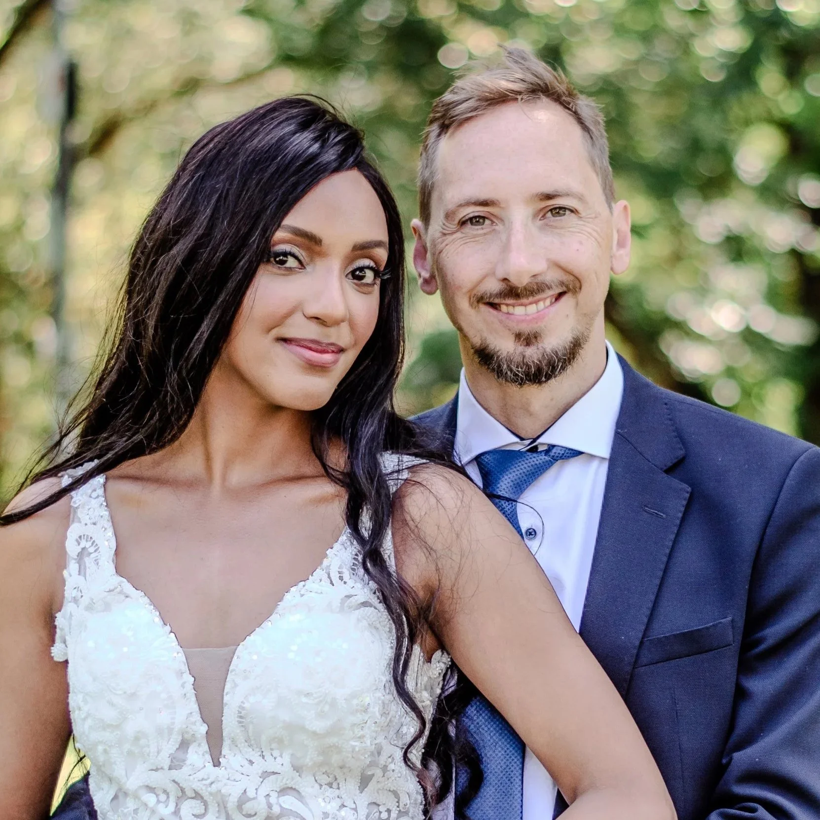 Ein Brautpaar bei einem Fotoshooting draußen, die Frau trägt ein weißes Hochzeitskleid und der Mann einen dunklen Anzug, umgeben von grünen Bäumen.