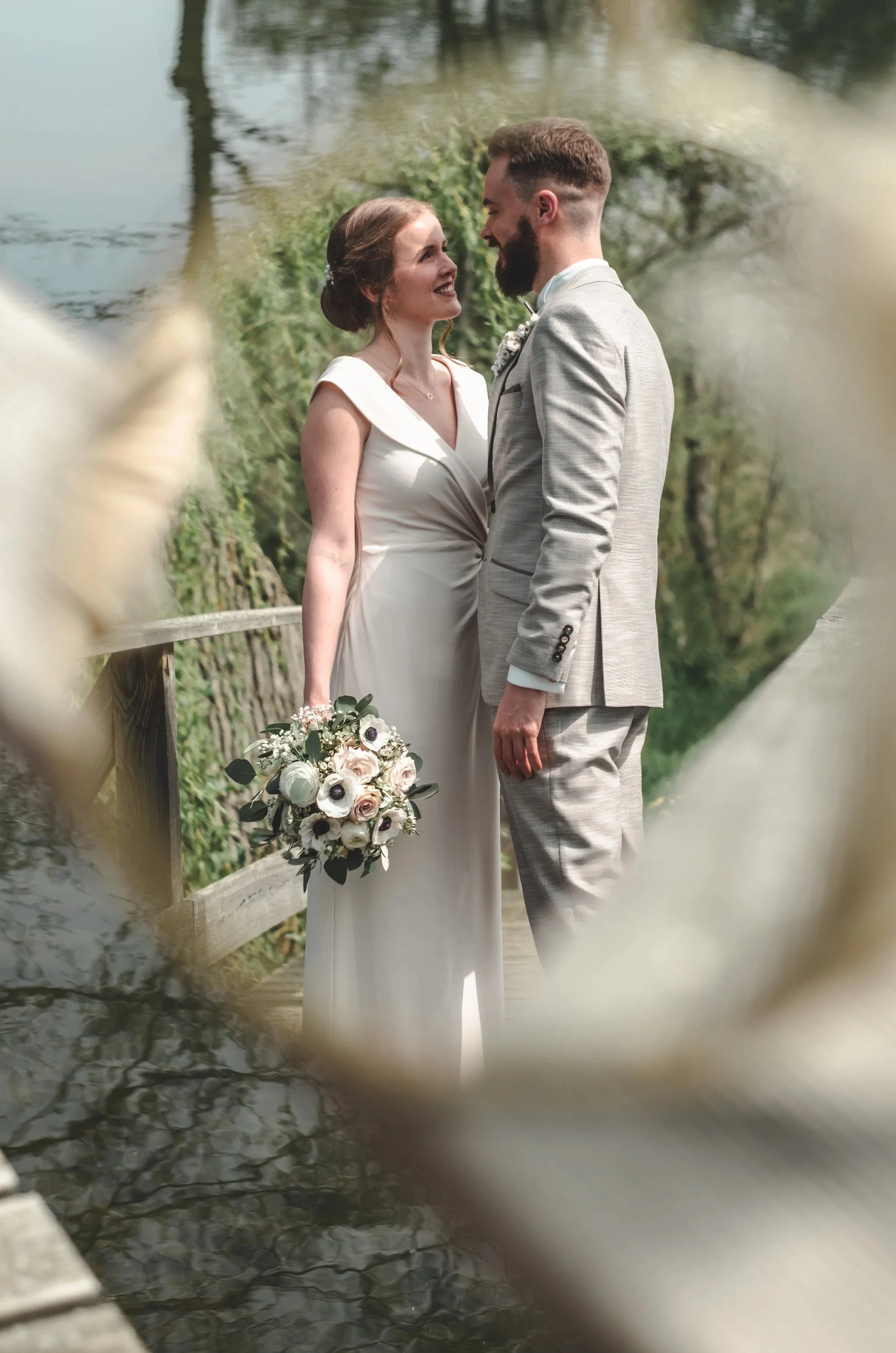Ein glückliches Brautpaar bei einer Hochzeit im Freien, die Frau trägt ein weißes Kleid und hält einen Blumenstrauß, der Mann trägt einen hellgrauen Anzug. Sie stehen sich gegenüber und lächeln sich an, im Hintergrund sind Bäume und Wasser zu sehen.