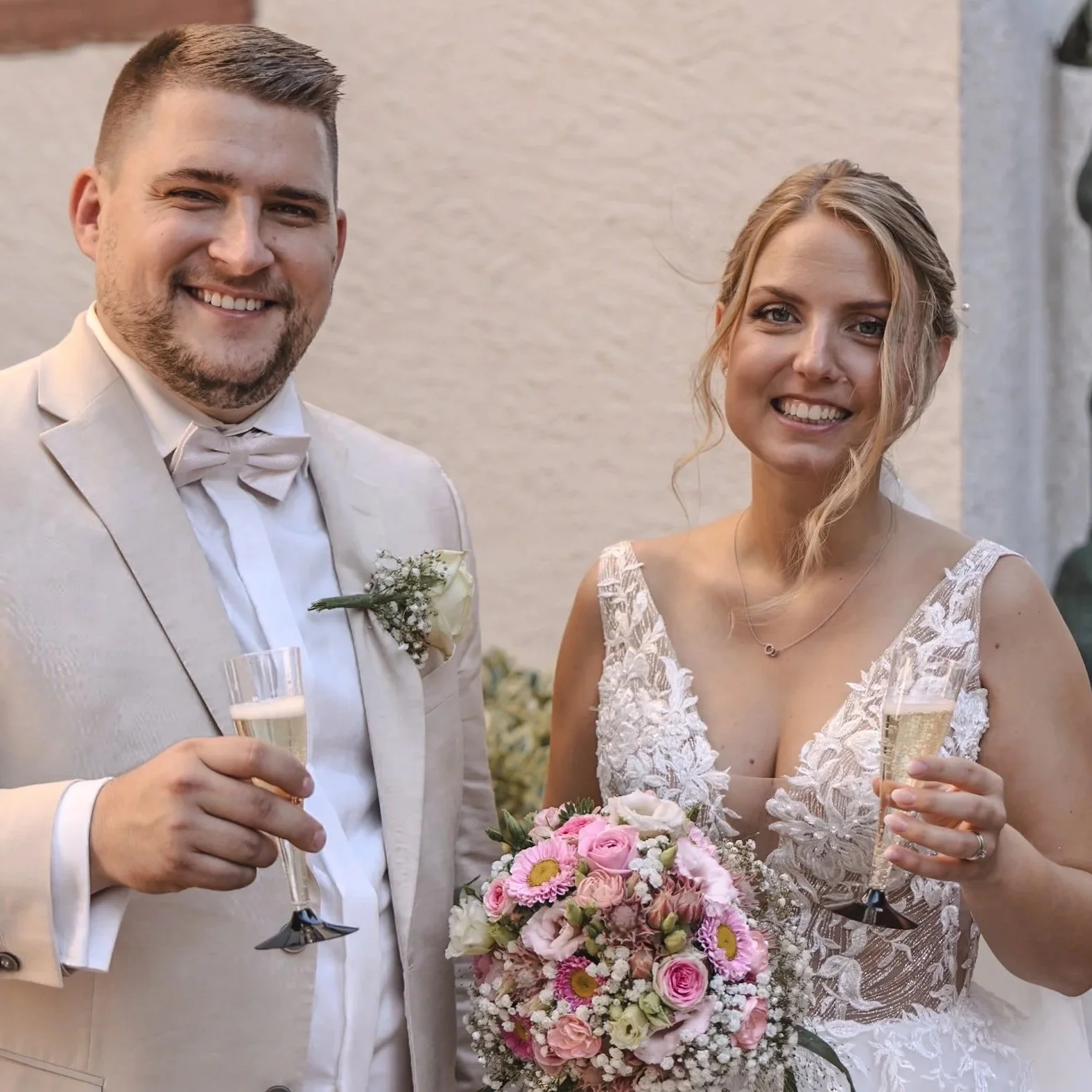 Ein frisch vermähltes Paar bei einer Hochzeit, beide lachend und in festlicher Kleidung, mit einem Blumenstrauß und Sektglas in der Hand.