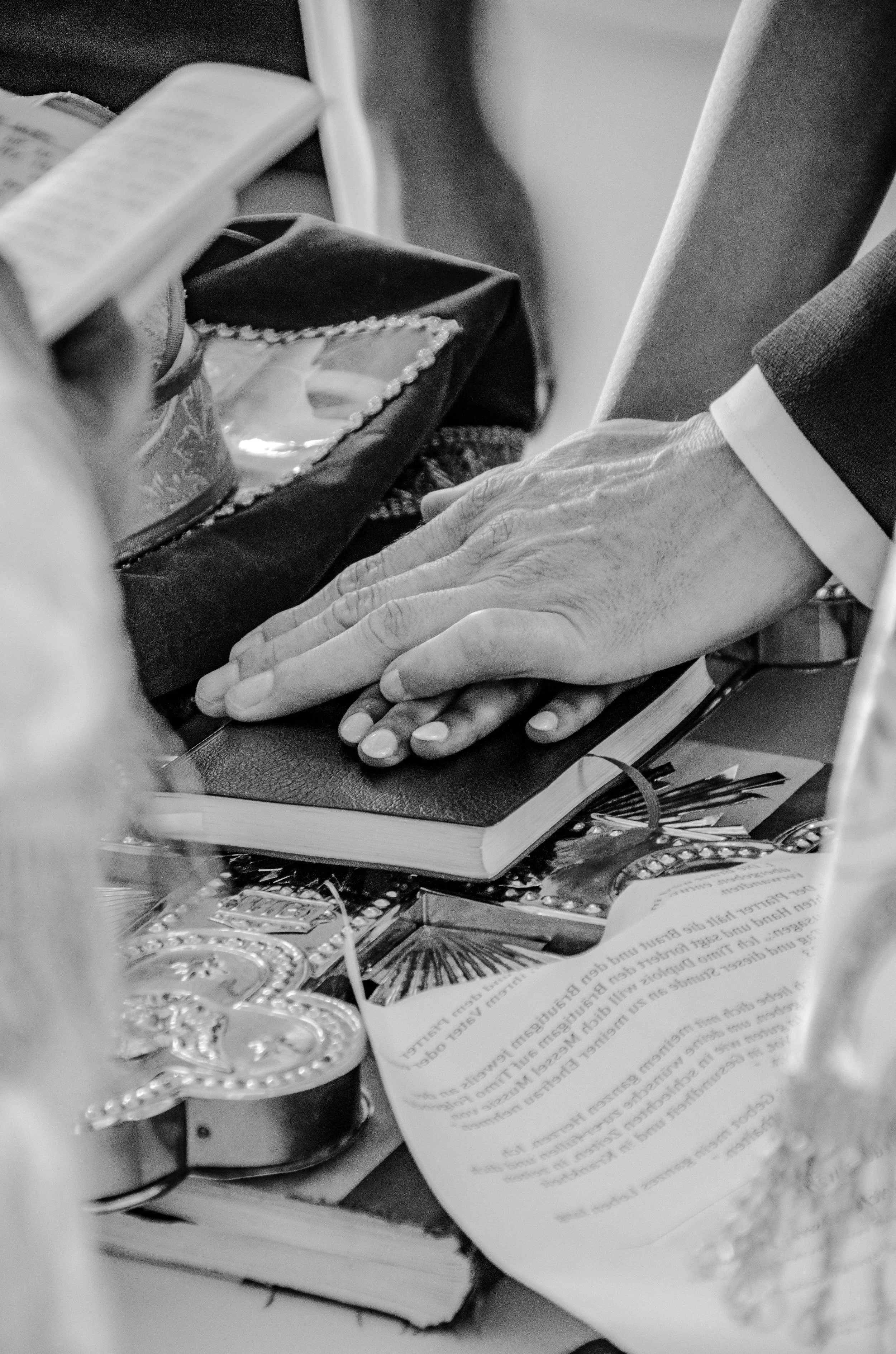 Nahaufnahme der Eheschliessung, das auf einem Tisch liegt, im Hintergrund ein Buch, eine Person in einem Anzug mit weißem Hemd und schwarzer Jacke hält die Hand auf den Tisch in der Kirche