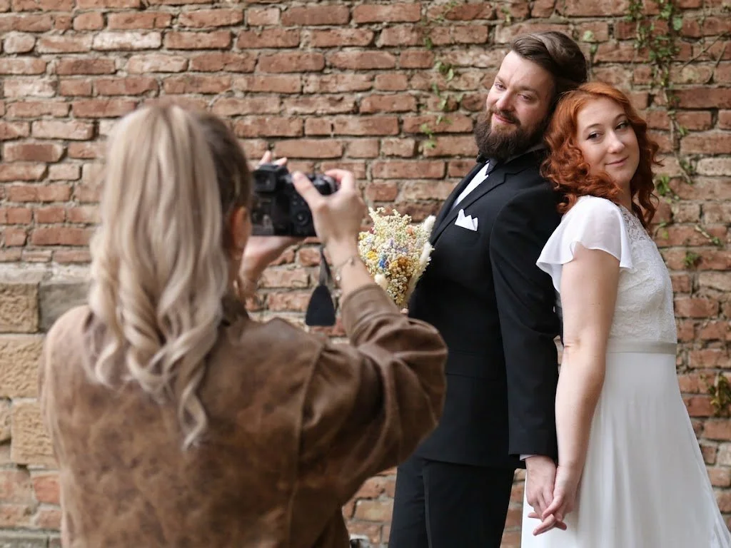 Ein Paar bei einer Hochzeit posaunt vor einer roten Backsteinmauer, während eine Fotografin sie fotografiert.