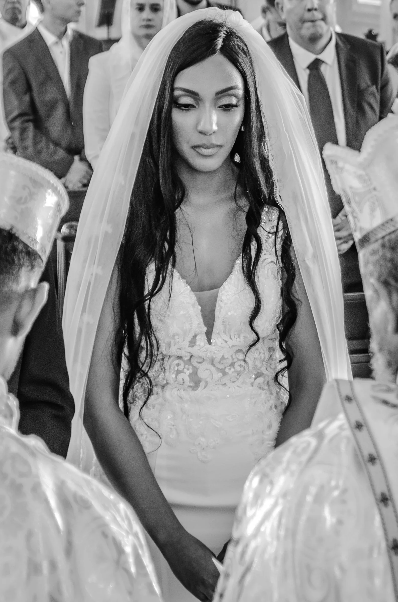 Eine Braut bei ihrer Zeremonie, umgeben von Gästen, in einem Hochzeitskleid und Schleier, in Schwarz-Weiß-Fotografie.