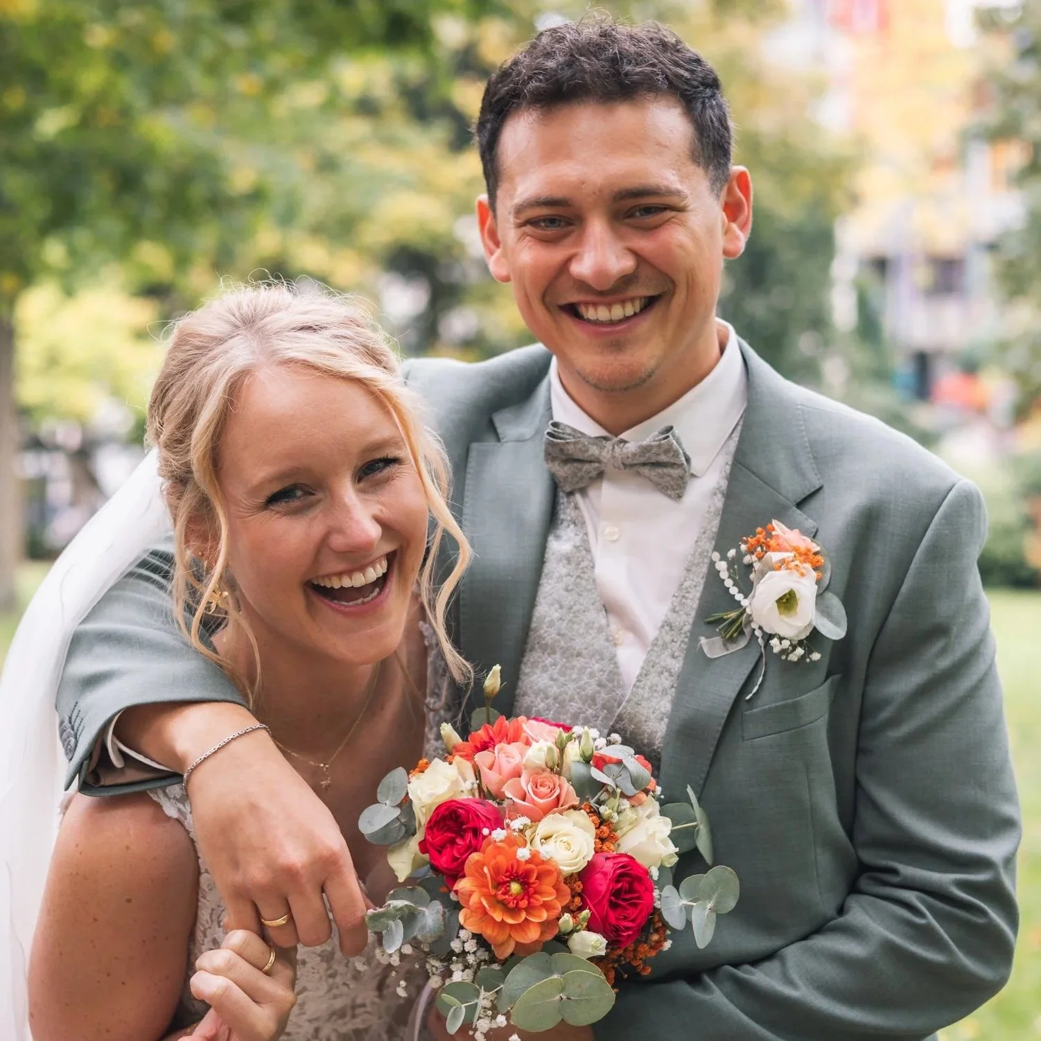 Ein glückliches Brautpaar lächelt bei einer Hochzeit im Freien, die Braut hält einen bunten Blumenstrauß, im Hintergrund Bäume und Gebäude.