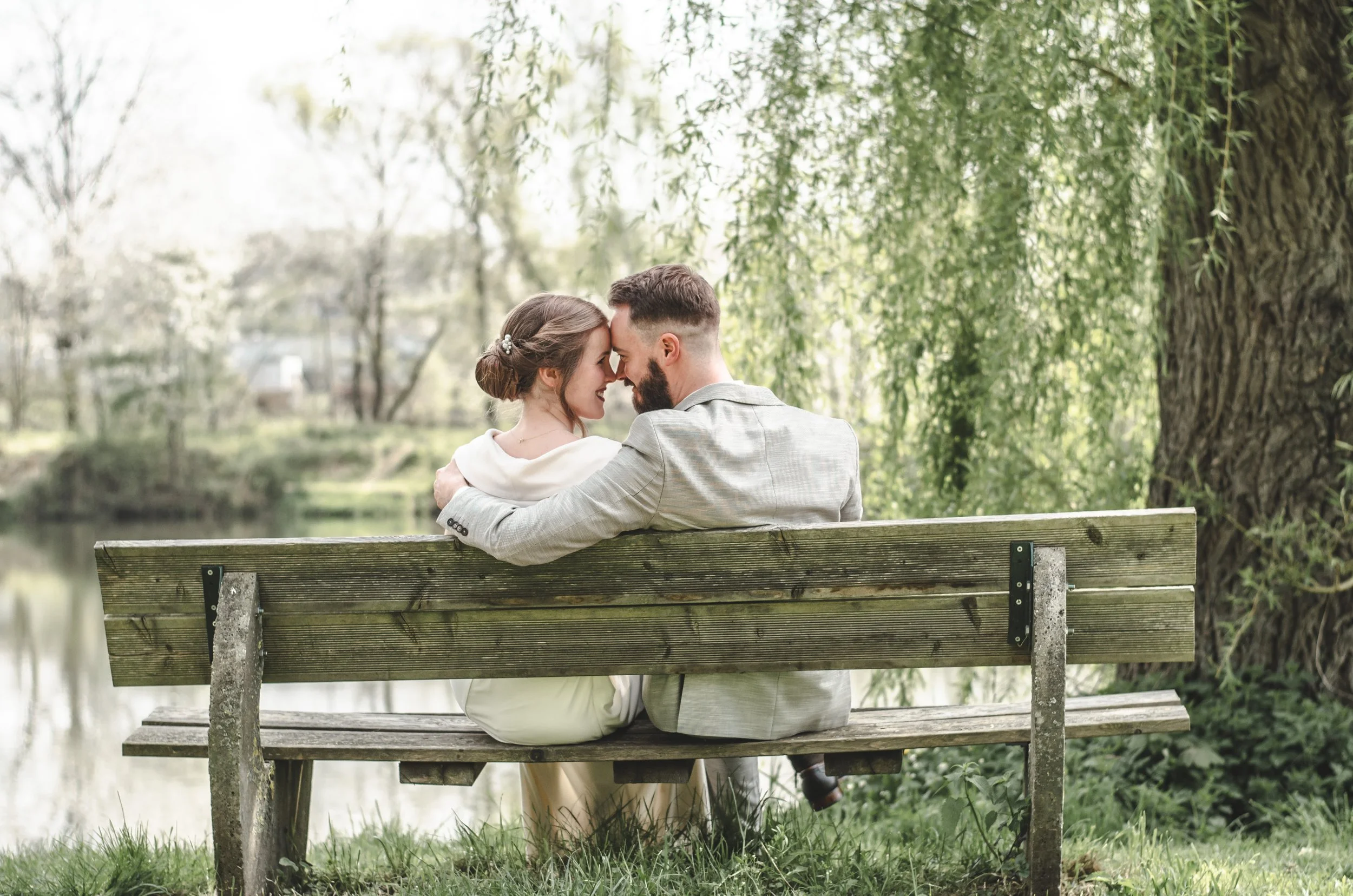 Ein verliebtes Hochzeitspaar sitzt auf einer Parkbank in einem parkähnlichen Gebiet mit Bäumen und einem Fluss im Hintergrund, sie berühren mit den Stirn und lächeln.