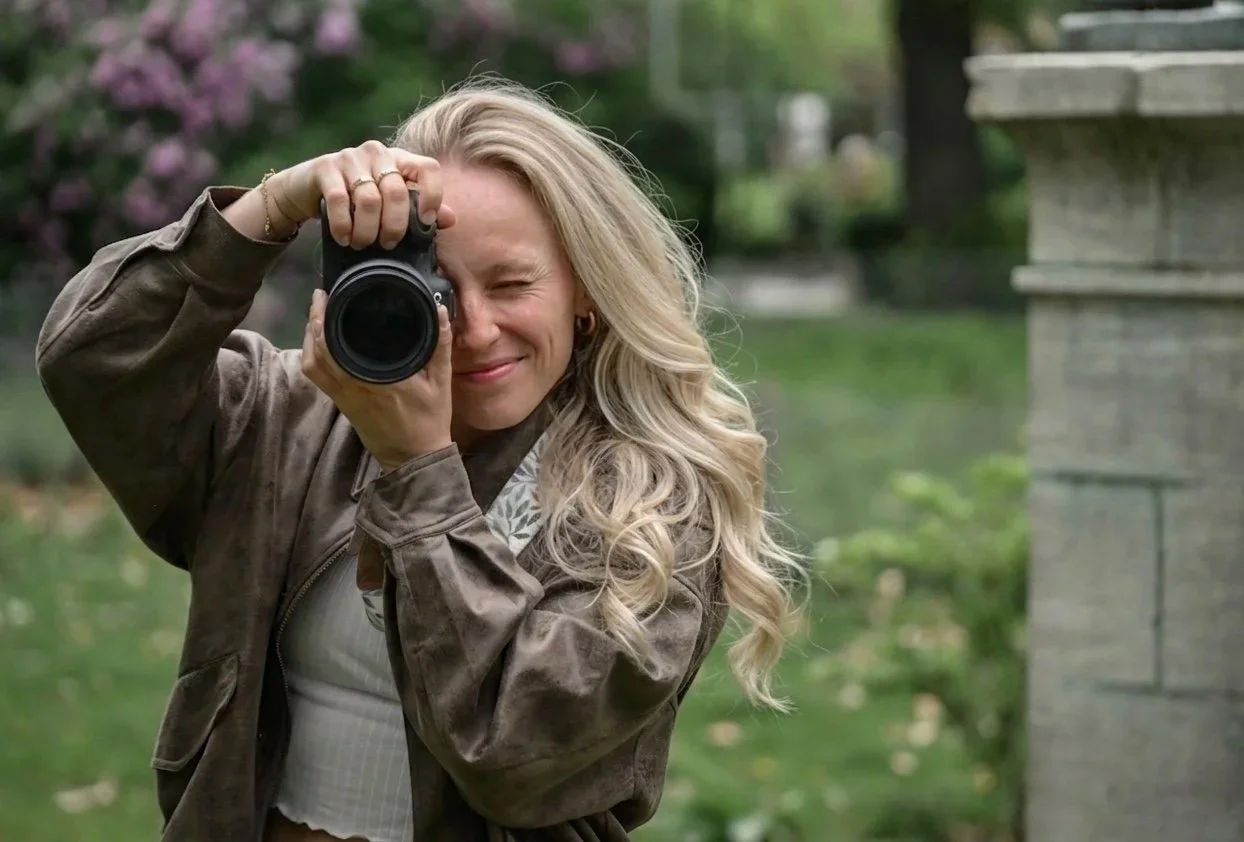 Eine lachende Frau mit langen, lockigen blonden Haaren hält eine Kamera vor einem Garten mit Bäumen und Blumen. Sie posiert beim Fotografieren.