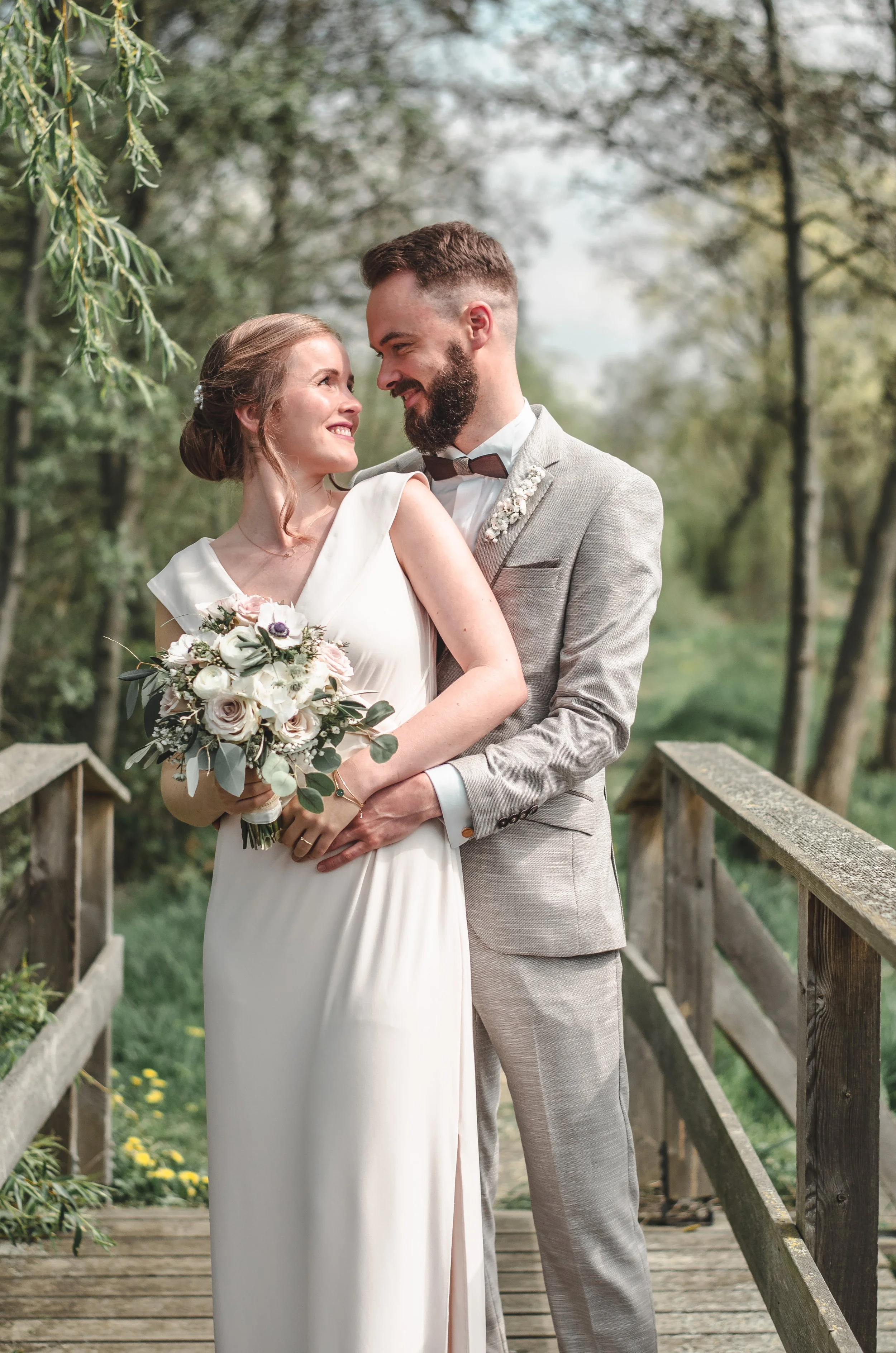 Ein heiratendes Paar auf einer Holzbrücke im Grünen, die Braut in weißem Kleid hält einen Brautstrauß, der Bräutigam in grauem Anzug, sie lächeln sich verliebt an.
