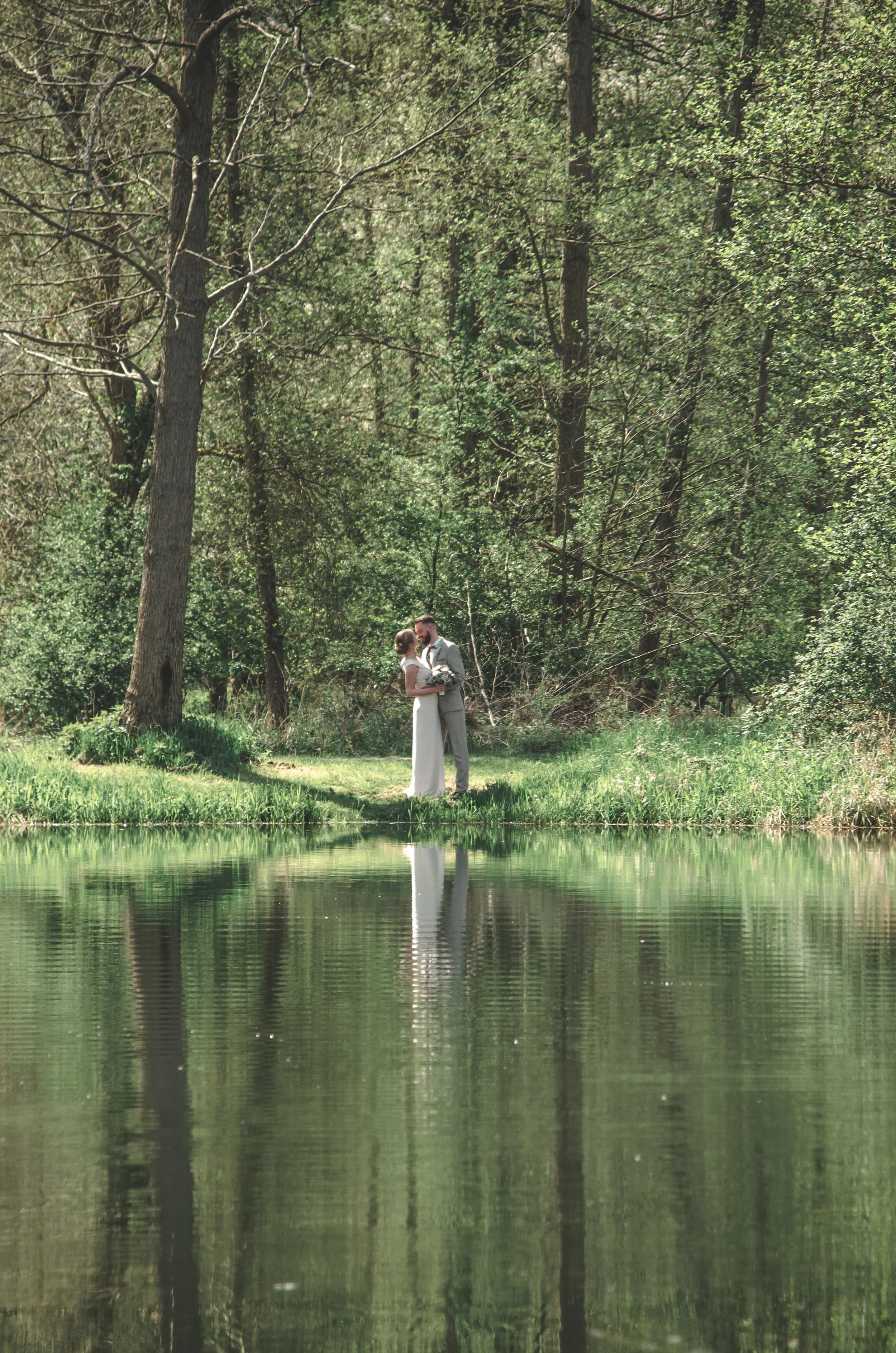 Ein Paar steht sich in einem grünen Wald bei Sonnenlicht gegenüber und küsst sich, während sich im Wasser die Spiegelung des Paares und der Bäume widerspiegelt.