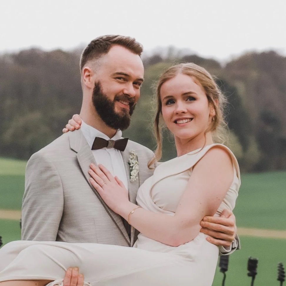 Ein junges, glückliches Brautpaar bei der Hochzeit im Freien, der Bräutigam trägt einen hellgrauen Anzug mit Fliege, die Braut trägt ein elegantes, cremefarbenes Kleid.