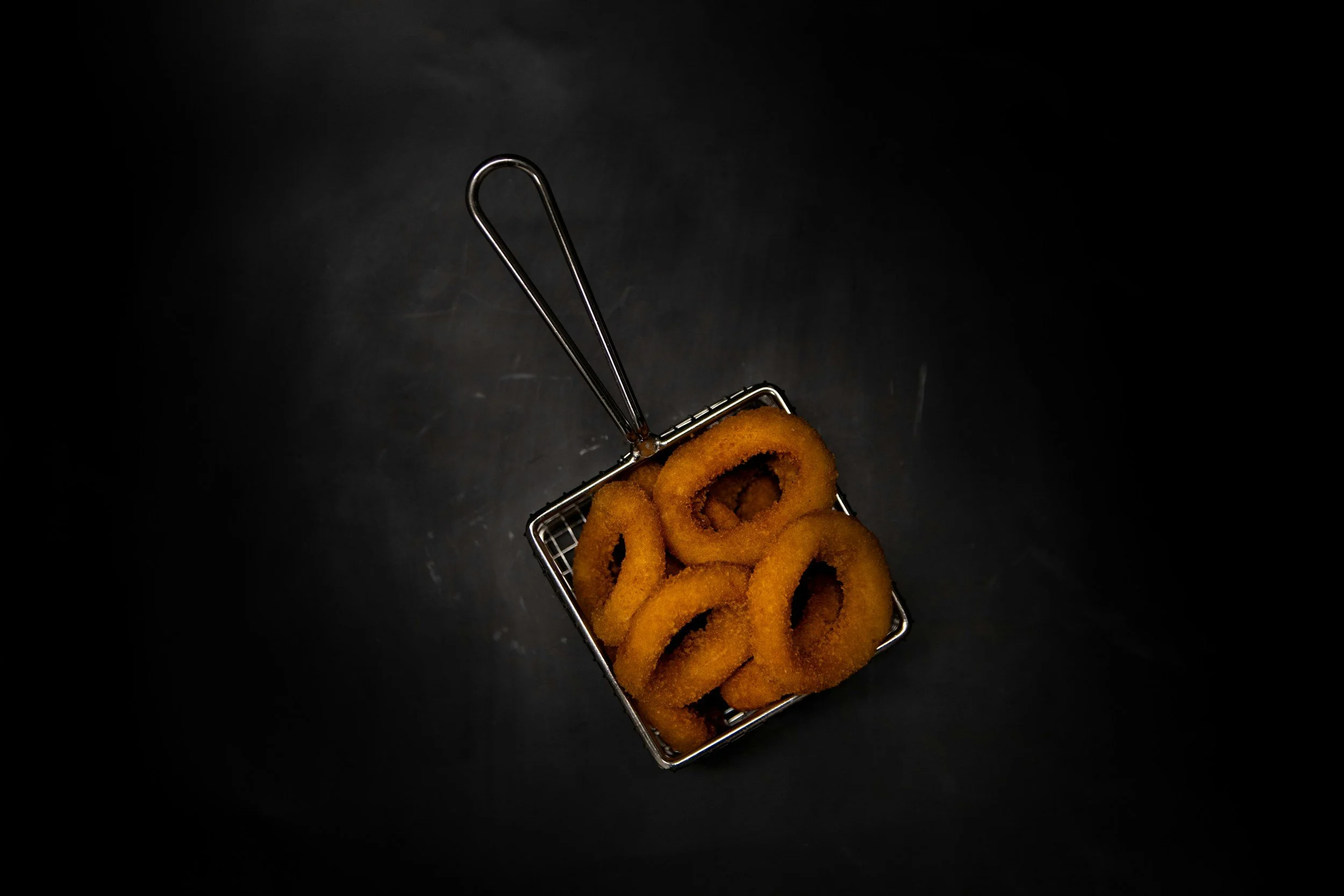 Golden-brown onion rings in a small metallic basket against a dark background.