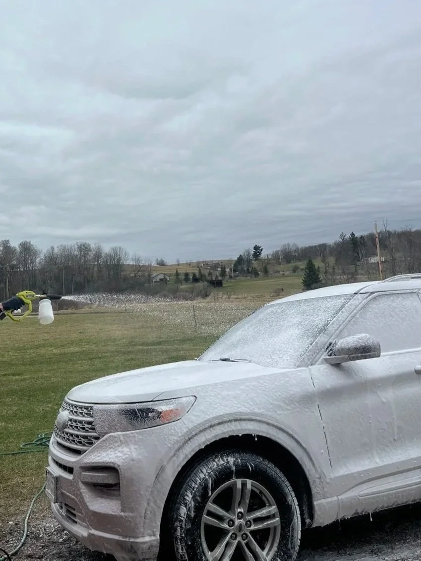 A silver SUV is being washed with foam soap outdoors on a cloudy day. A person is using a spray nozzle to rinse the vehicle.