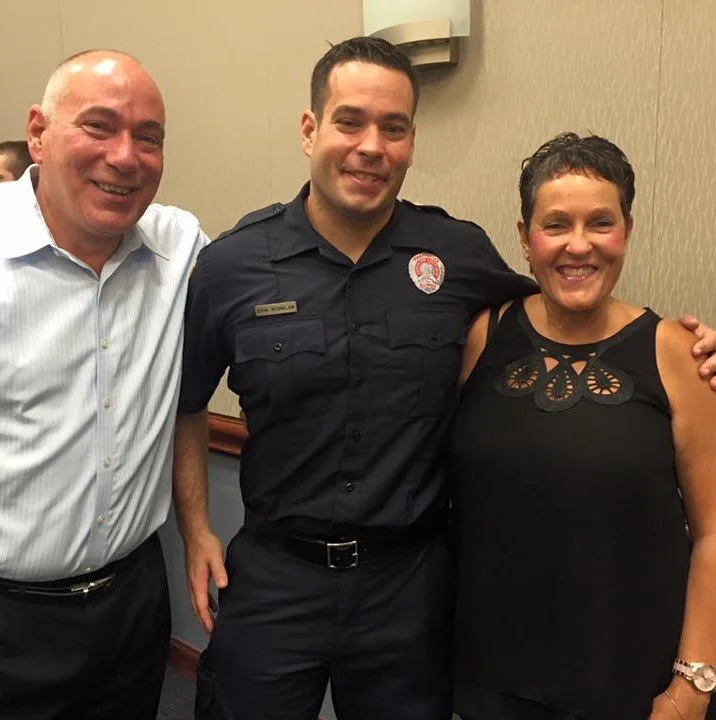 Three people standing together indoors, smiling, with a beige wall and a wall-mounted light fixture behind them. The person in the middle is a firefighter in uniform.