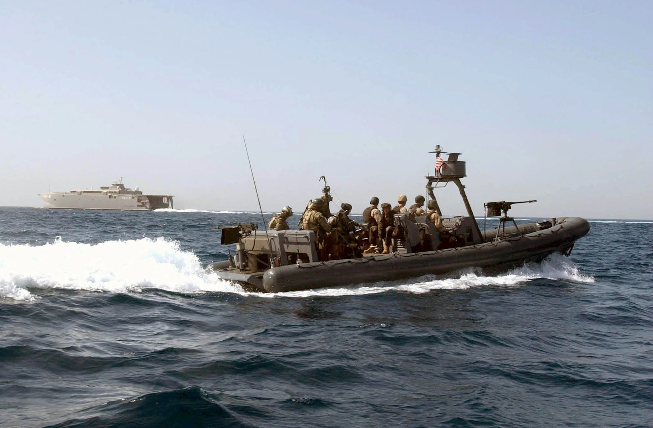 Military personnel on a rigid inflatable boat with machine guns, sailing in the ocean with a large gray aircraft carrier in the background.