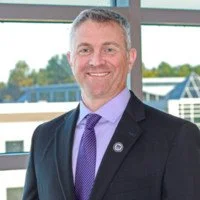 A man in a dark suit, purple tie, and light blue shirt standing outdoors with a modern building and trees in the background.