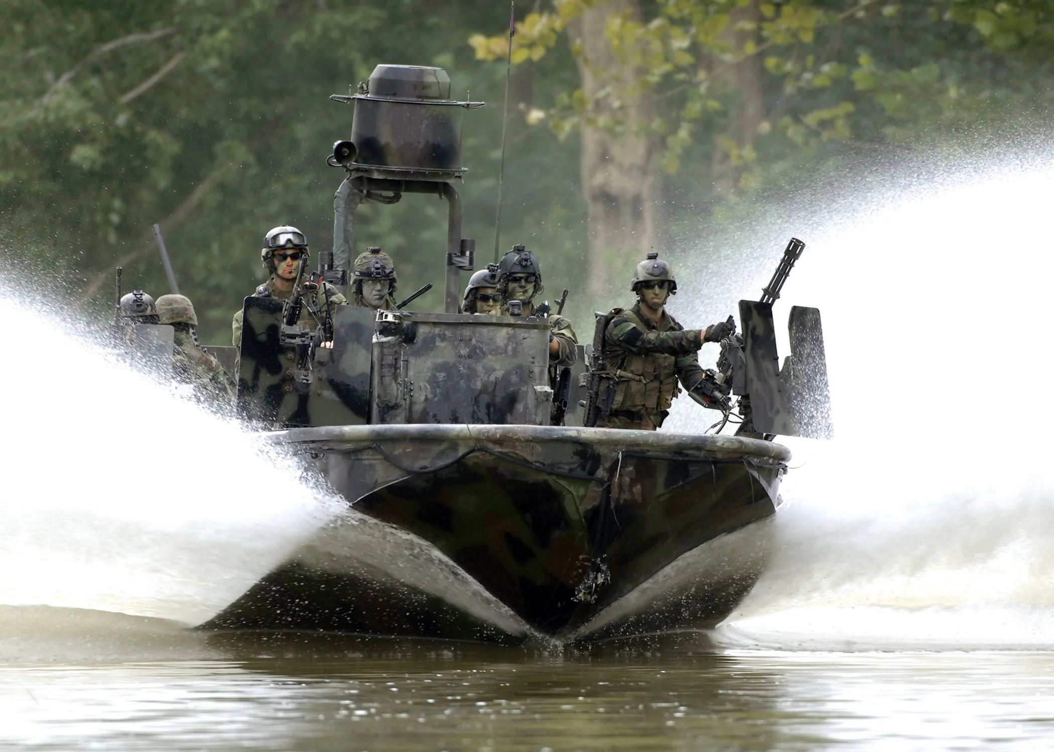 Military personnel on a boat moving at high speed through a water body, wearing tactical gear and helmets.