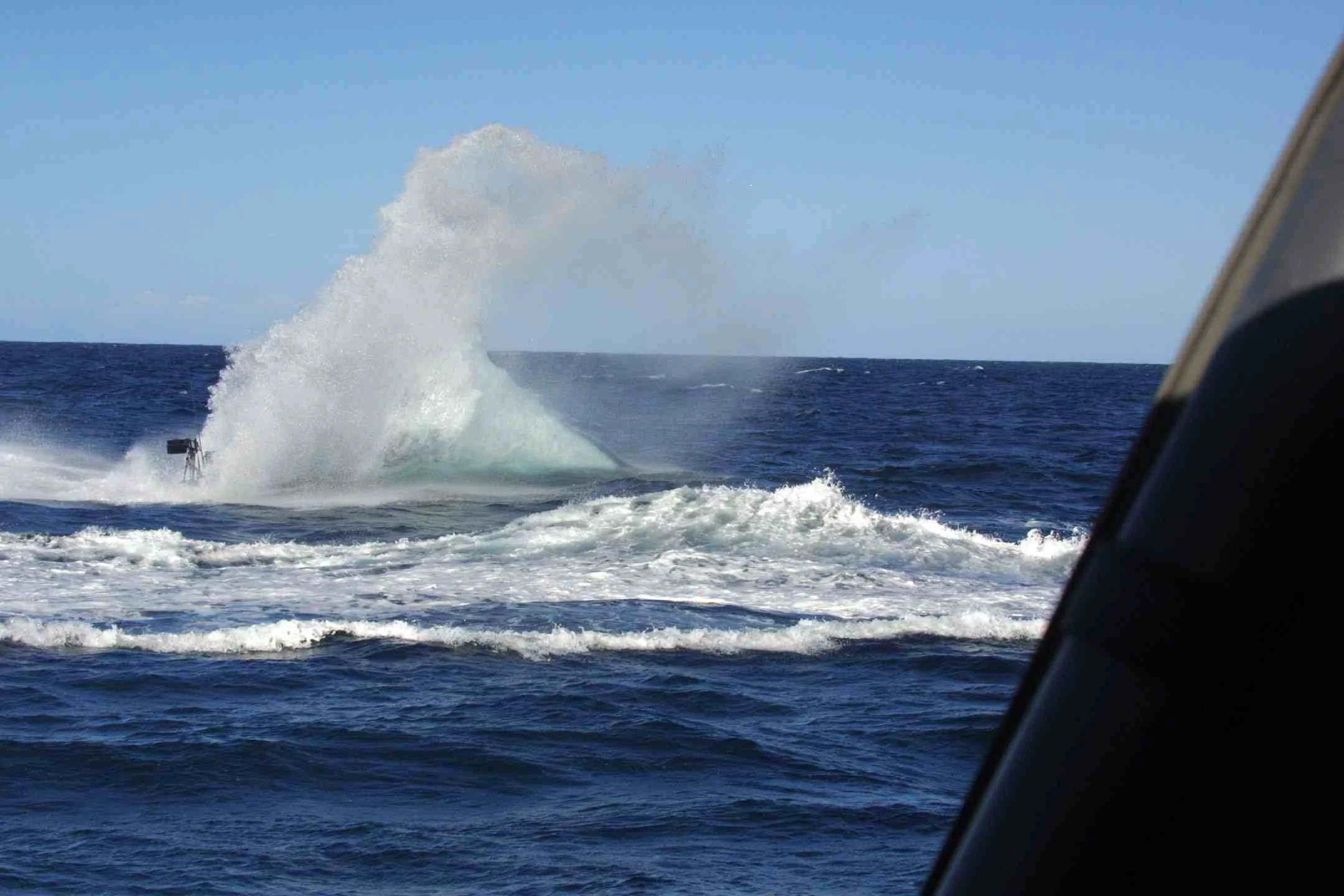 A boat moving at high speed on the ocean, creating a large spray of water in its wake, with a clear blue sky in the background.