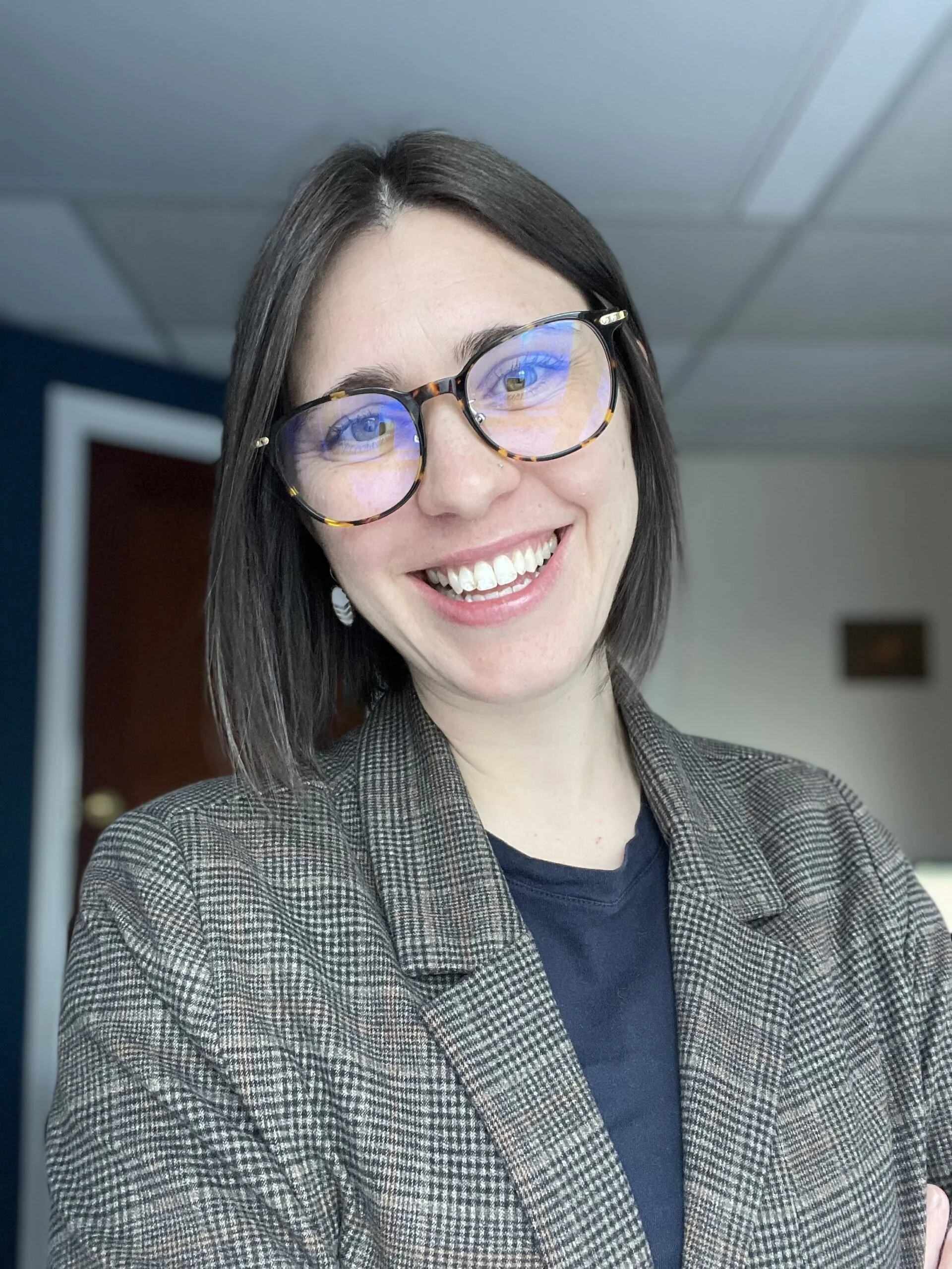 A woman with dark hair, wearing glasses with tortoiseshell frames, smiling and showing her teeth, dressed in a checked blazer over a dark shirt, in an indoor setting.
