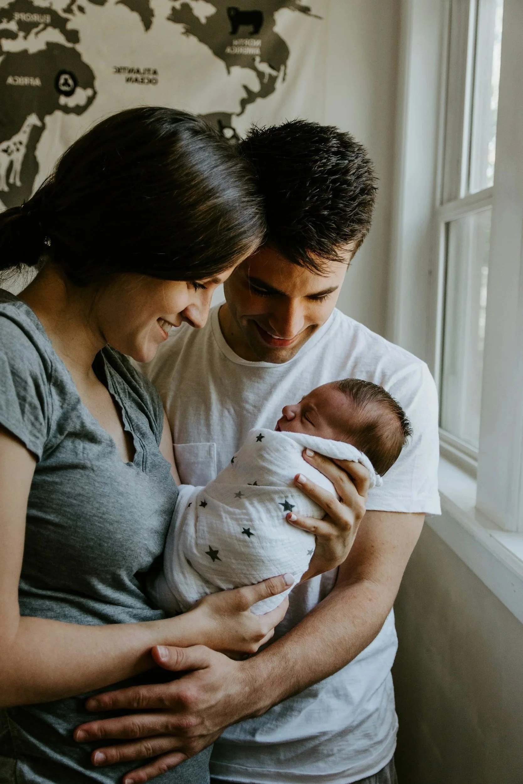 A family holding a newborn baby, smiling and looking at the baby lovingly in a cozy room with a world map on the wall.