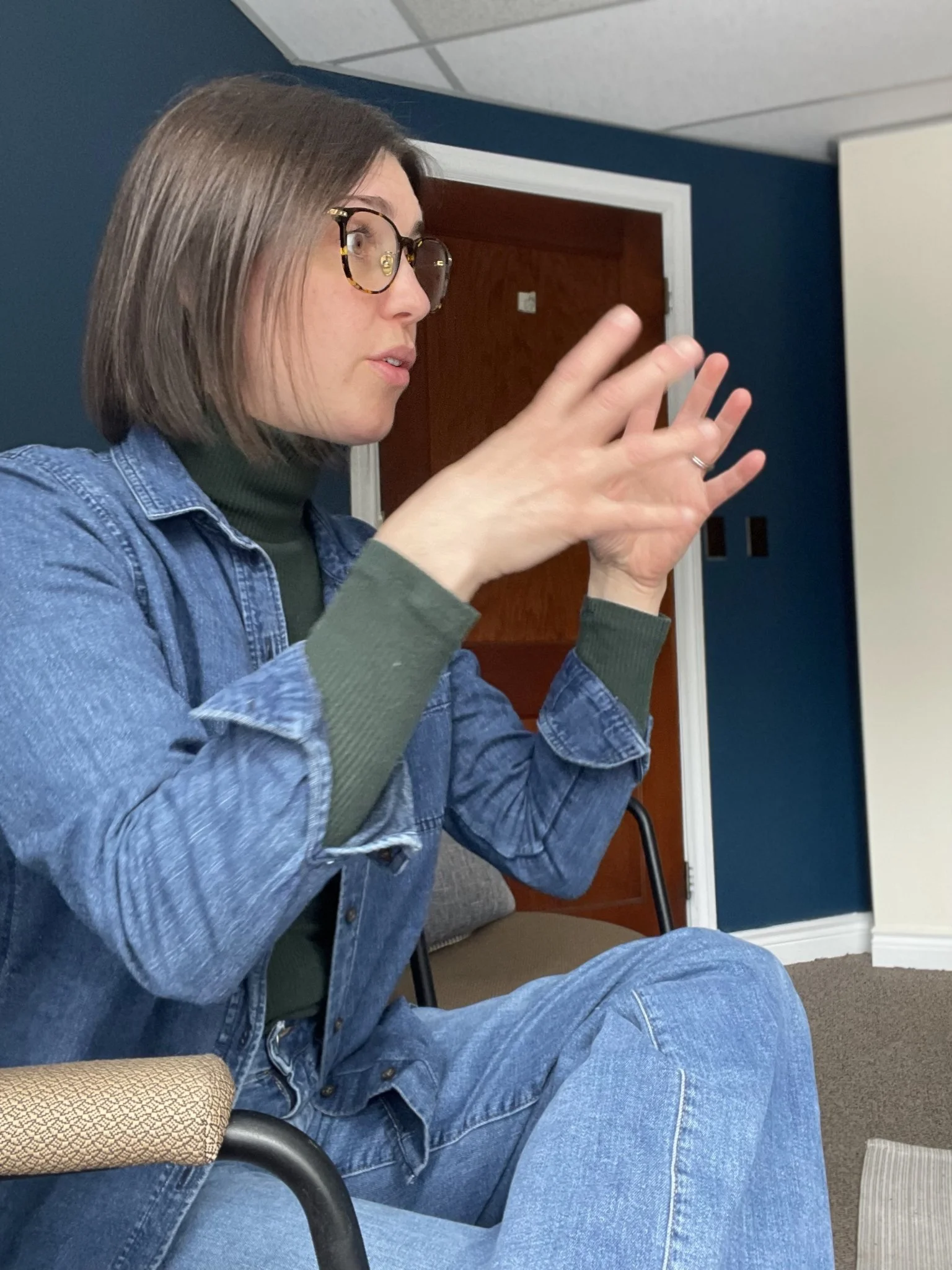 A woman with shoulder-length brown hair, wearing glasses, a green turtleneck, and a denim jacket, is seated and appears to be speaking or explaining something, with her hands raised.