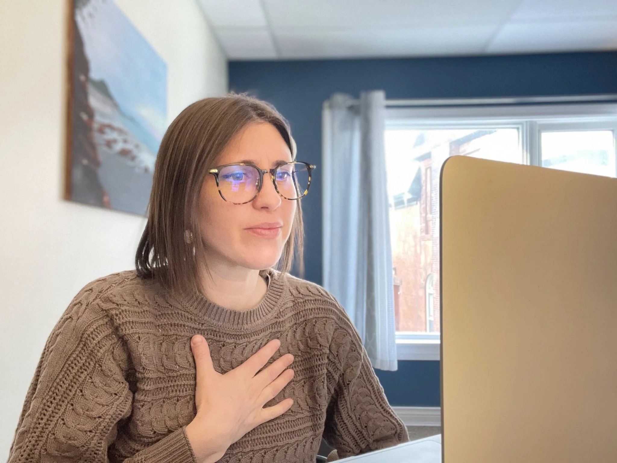 A woman with brown hair and glasses sitting at a desk with one hand over her chest in a room with a blue wall, a window with white curtains, and artwork on the wall.