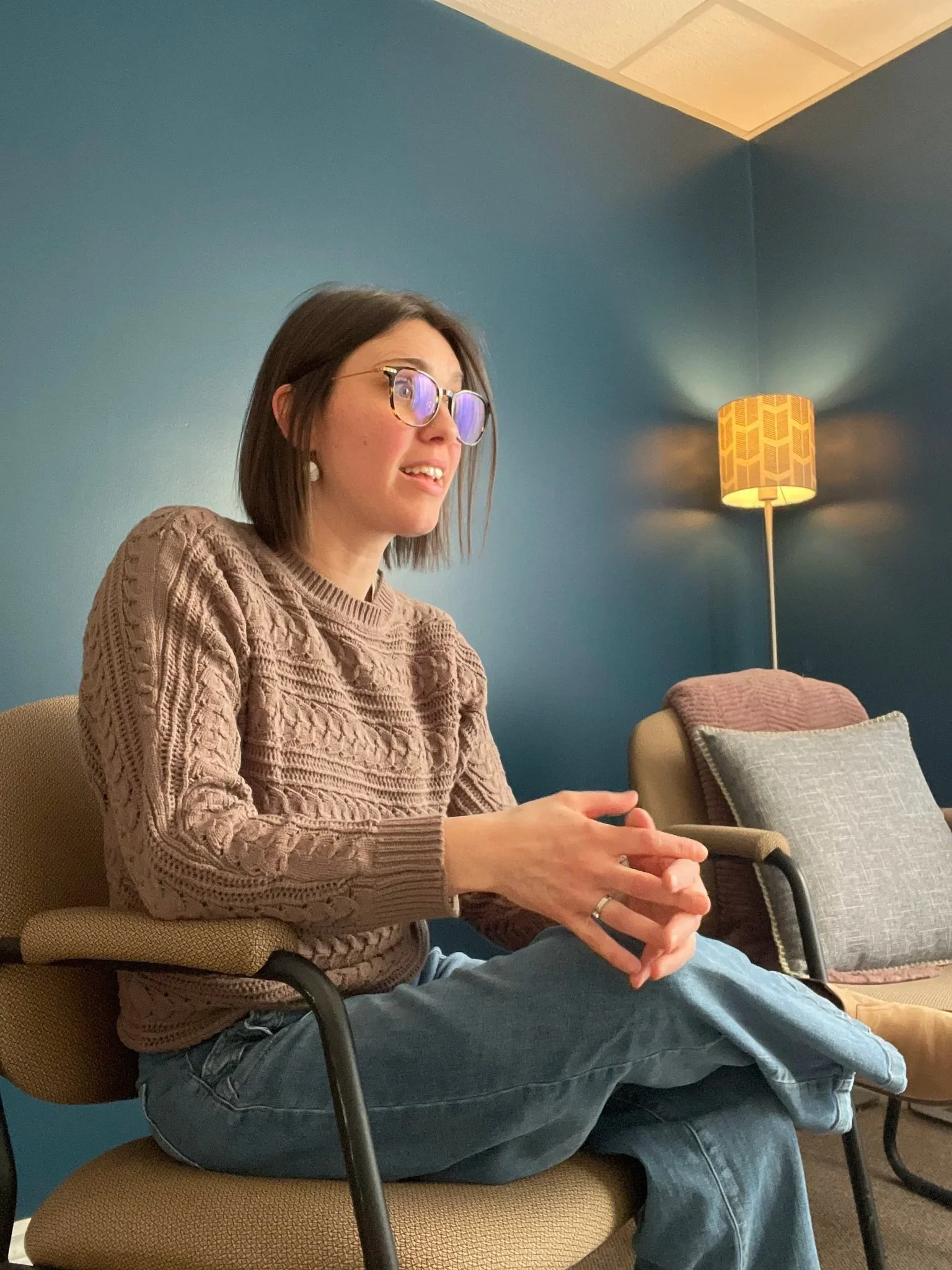 A woman wearing glasses and a beige sweater sitting in a waiting room, smiling and looking to her right. There is a blue wall, a floor lamp, and a beige chair with a pillow in the background.