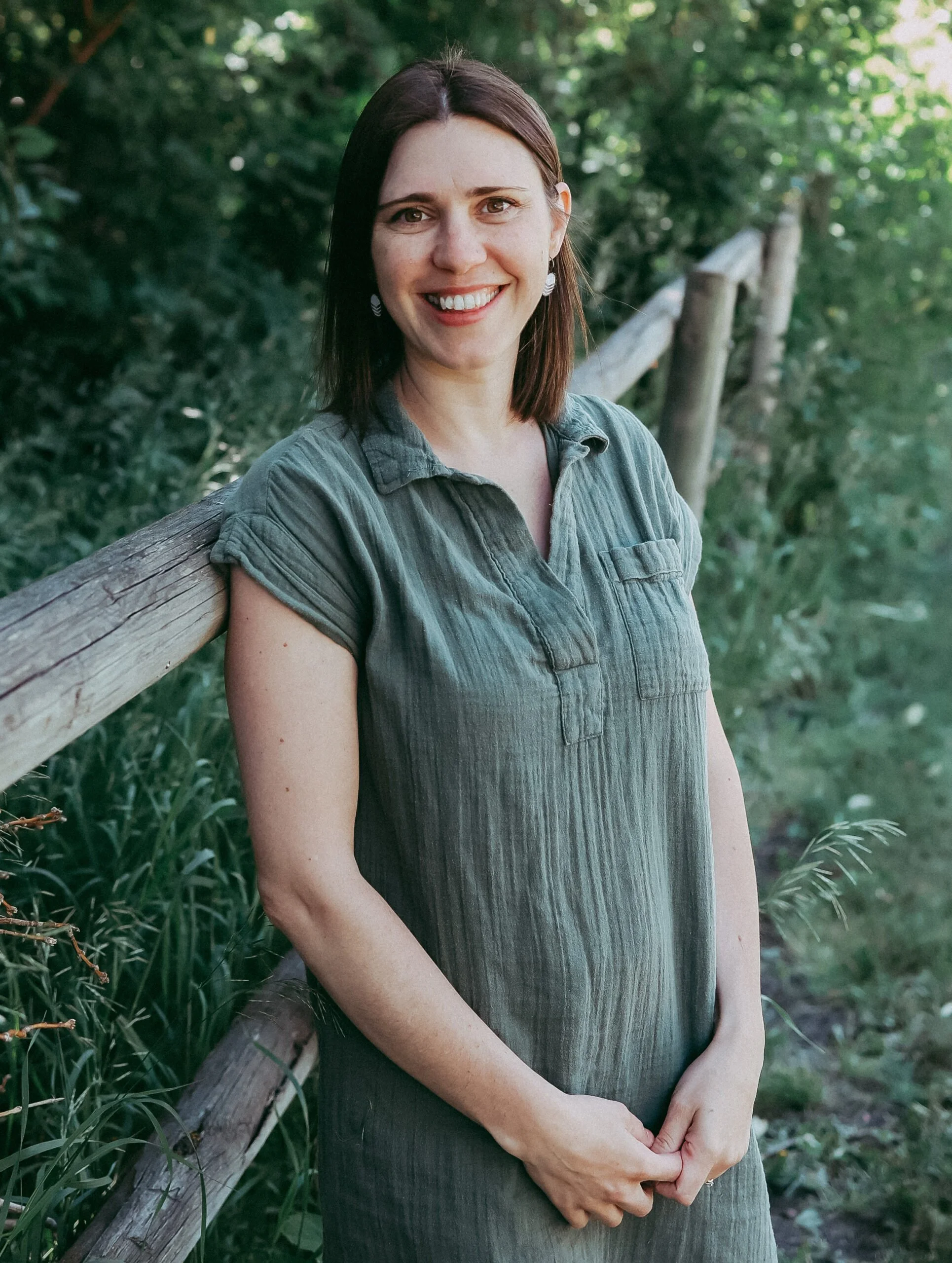 A woman with dark brown hair, wearing a gray shirt, stands outdoors leaning against a wooden railing with green foliage in the background.