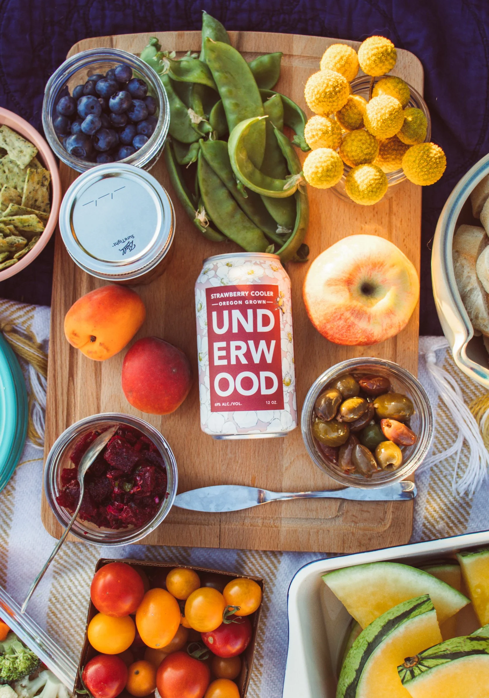 Assorted fresh fruits, vegetables, and snacks on a wooden platter, including blueberries, snap peas, yellow and red cherry tomatoes, an apple, a peach, a can of strawberry cooler, and bowls of olives, beet relish, and sliced melons.