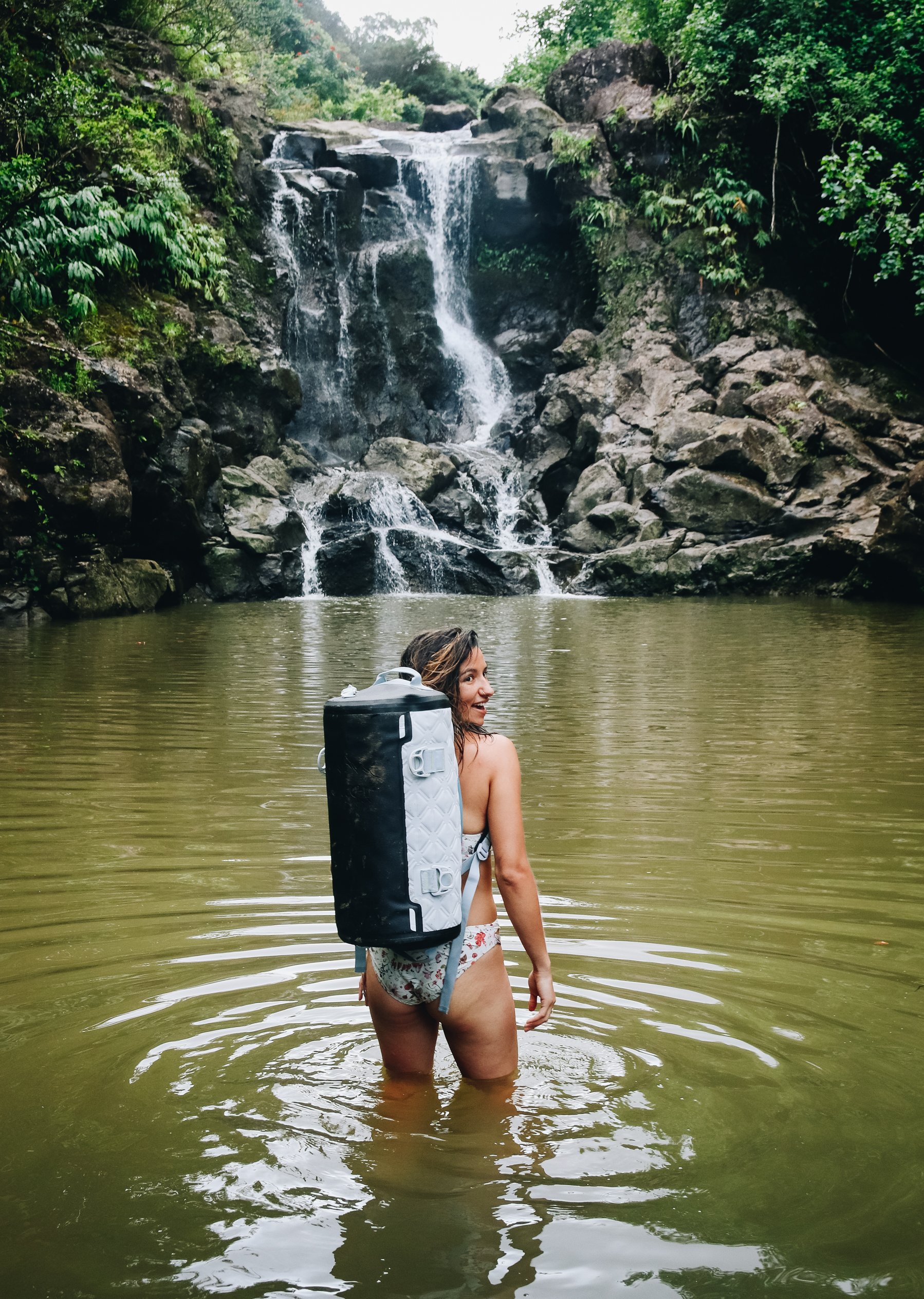A woman with a backpack standing in a river near a waterfall surrounded by greenery.
