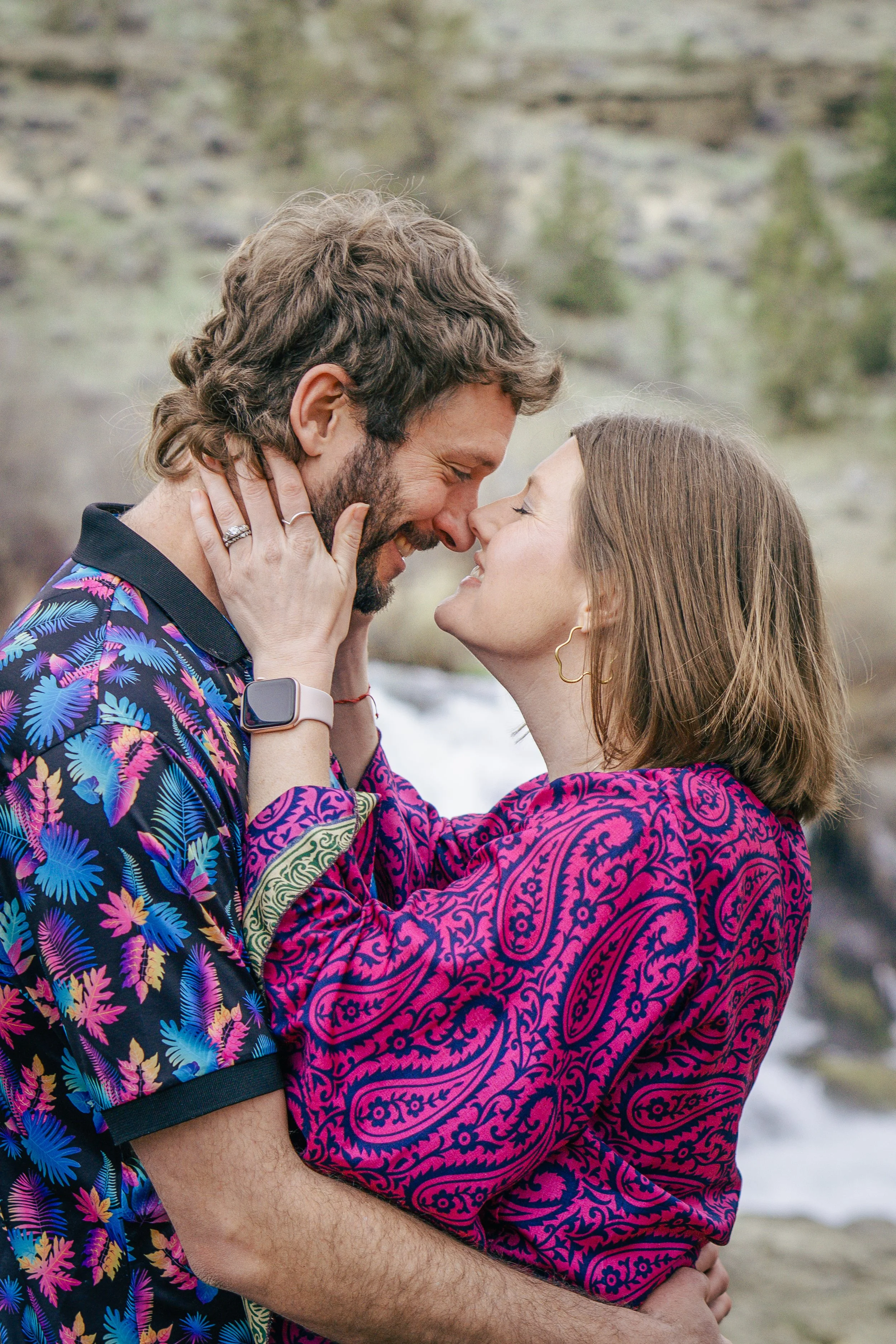 A couple embracing outdoors, close to a river, touching foreheads and smiling
