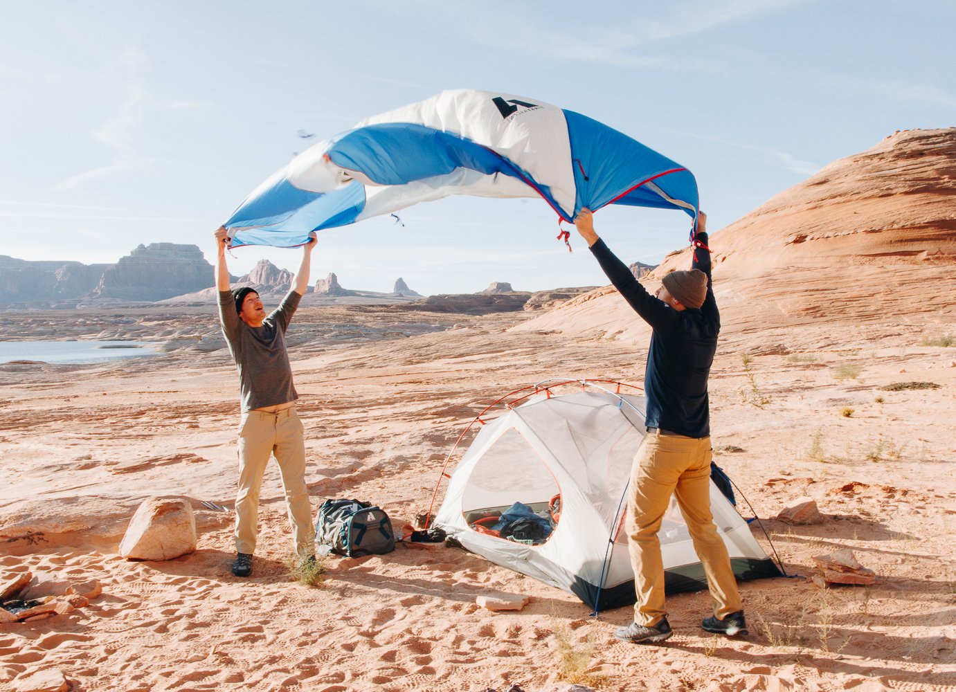 Two people set up a tent on a sandy desert landscape with rock formations and a body of water in the background, as they hold a deflated parachute or canopy above them.