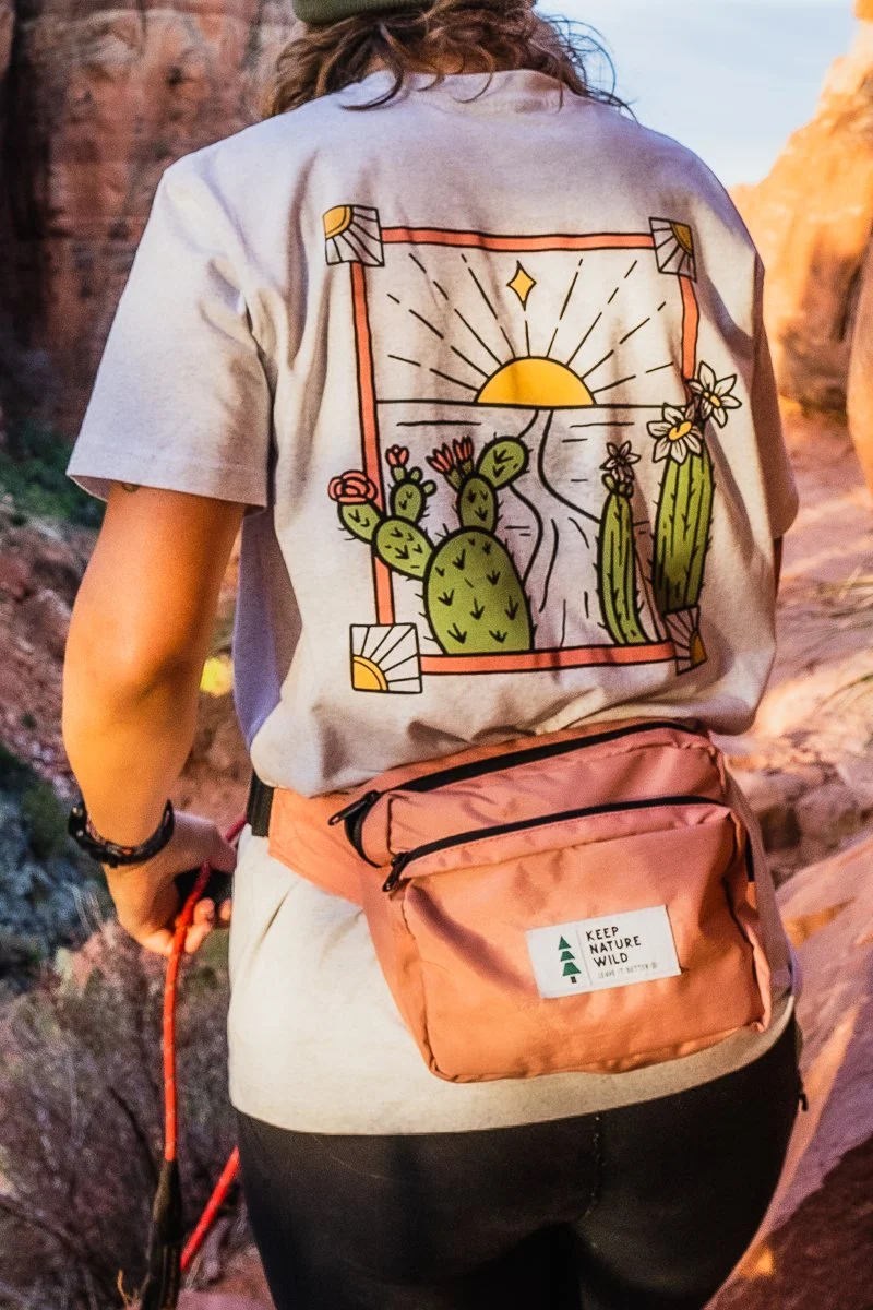 Back of a person wearing a gray t-shirt with a colorful cactus and sunset graphic, and a peach-colored waist pack with a "Keep Nature Wild" patch, hiking in a canyon at sunset.