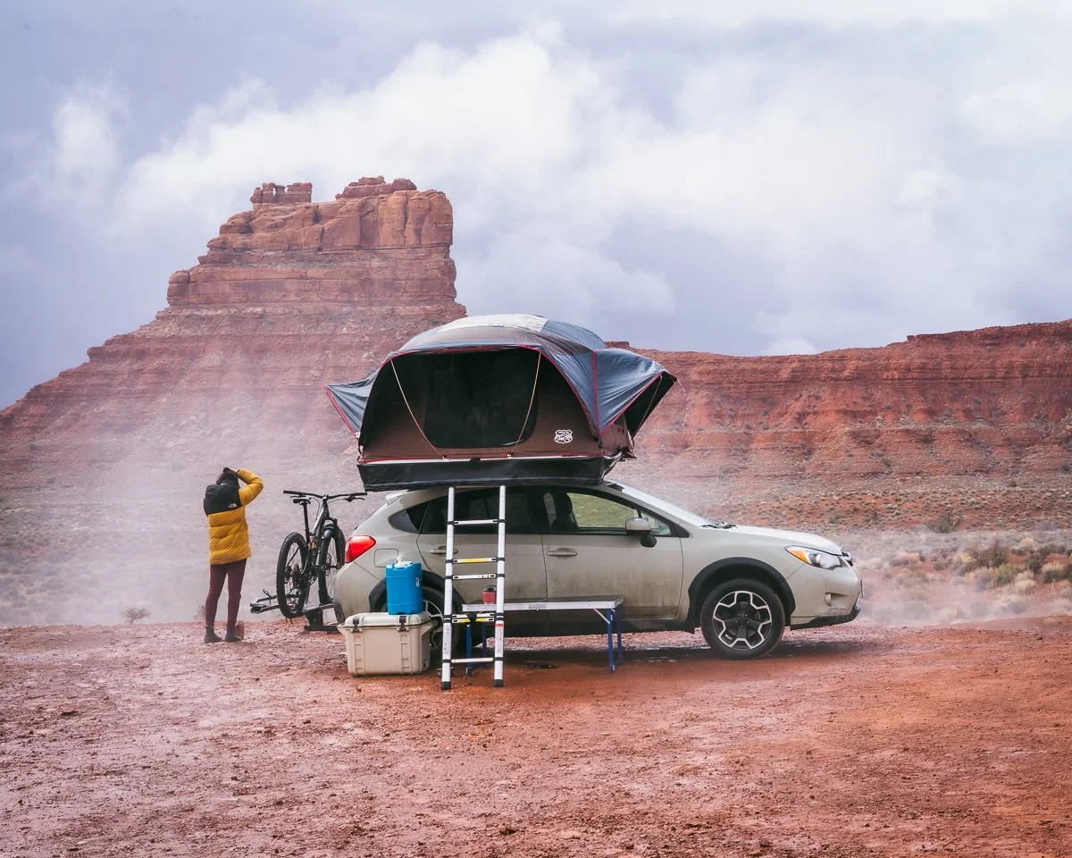 A car with a roof tent and bike attached, parked in a desert landscape with red rock formations in the background, person in yellow jacket standing nearby.