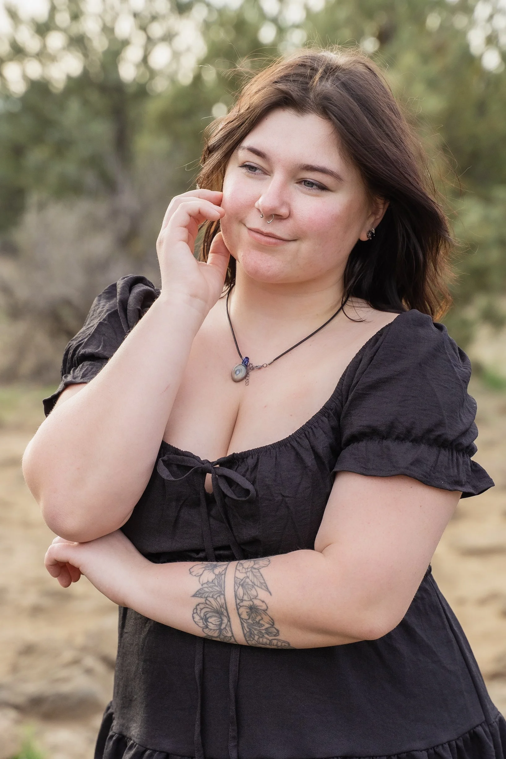 A young woman with shoulder-length brown hair, tattoos on her left arm, wearing a black dress with puffed sleeves and a lace-up front, accessorized with a necklace and ear piercings, standing outdoors with trees in the background.