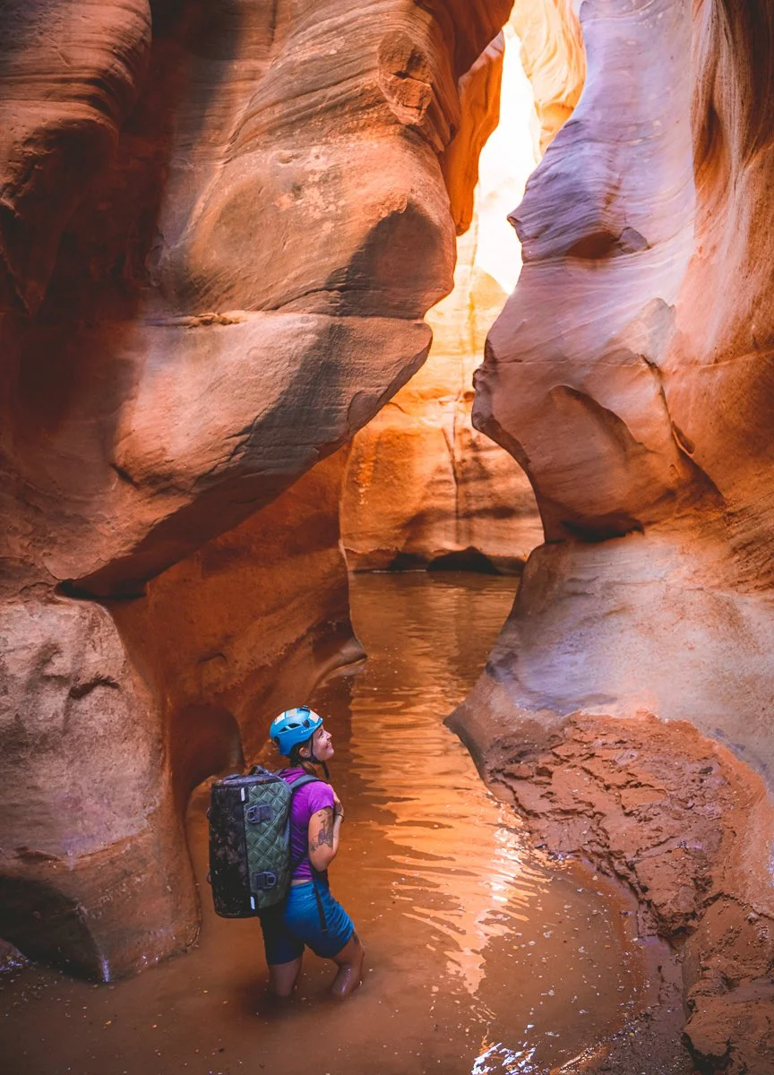 A woman standing in a narrow slot canyon with reddish-orange rock walls and water at her knees, wearing a blue helmet, purple shirt, and carrying a black backpack.