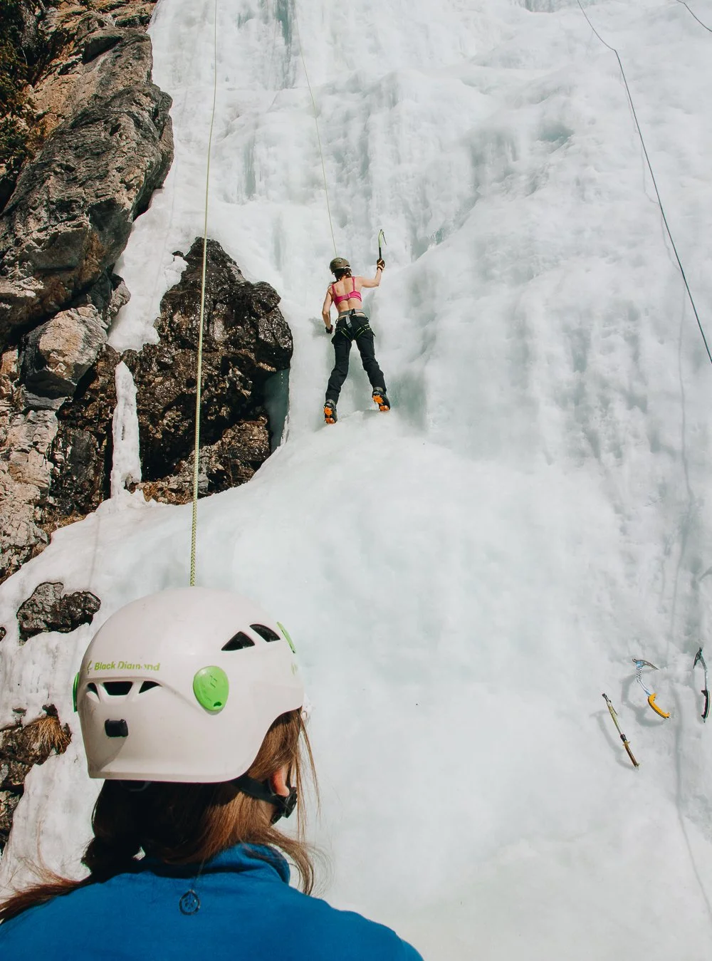 A person in climbing gear is ice climbing on a frozen waterfall, with a second person watching in the foreground wearing a helmet.